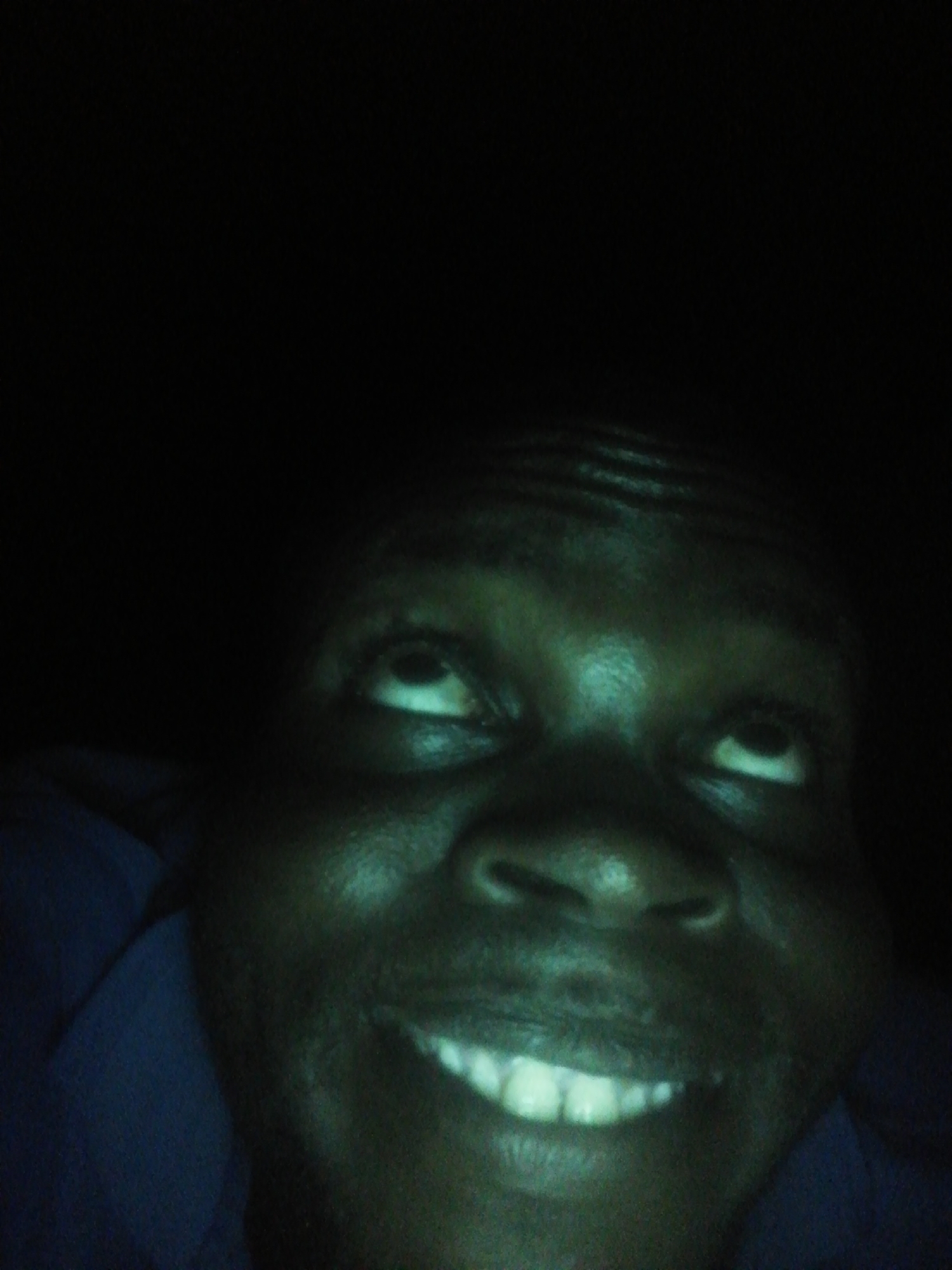 A close-up photograph shows a person with dark skin looking upwards and smiling. The lighting is dim, and a portion of a blue garment is visible in the lower left of the frame. The background is dark and indistinct. No information about the location or the city is available from the image.