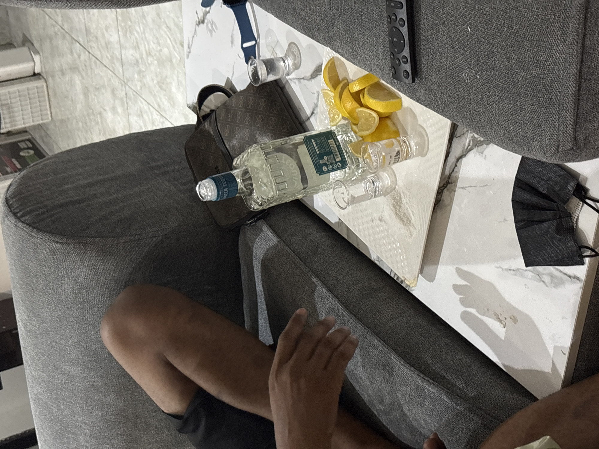 A photograph taken from an elevated perspective shows a portion of a person seated on a gray upholstered sofa. The individual’s legs and one hand are visible. A white marble-patterned side table sits next to the sofa, holding a clear bottle of what appears to be tequila, three small shot glasses, and a plate of lemon slices. There is also a Guess handbag and a black face mask on the table, as well as a blue sports watch and a black remote control. Part of a white storage unit can be seen in the background. The precise location in Iddo Tudunwada, Nigeria, cannot be determined from the image.