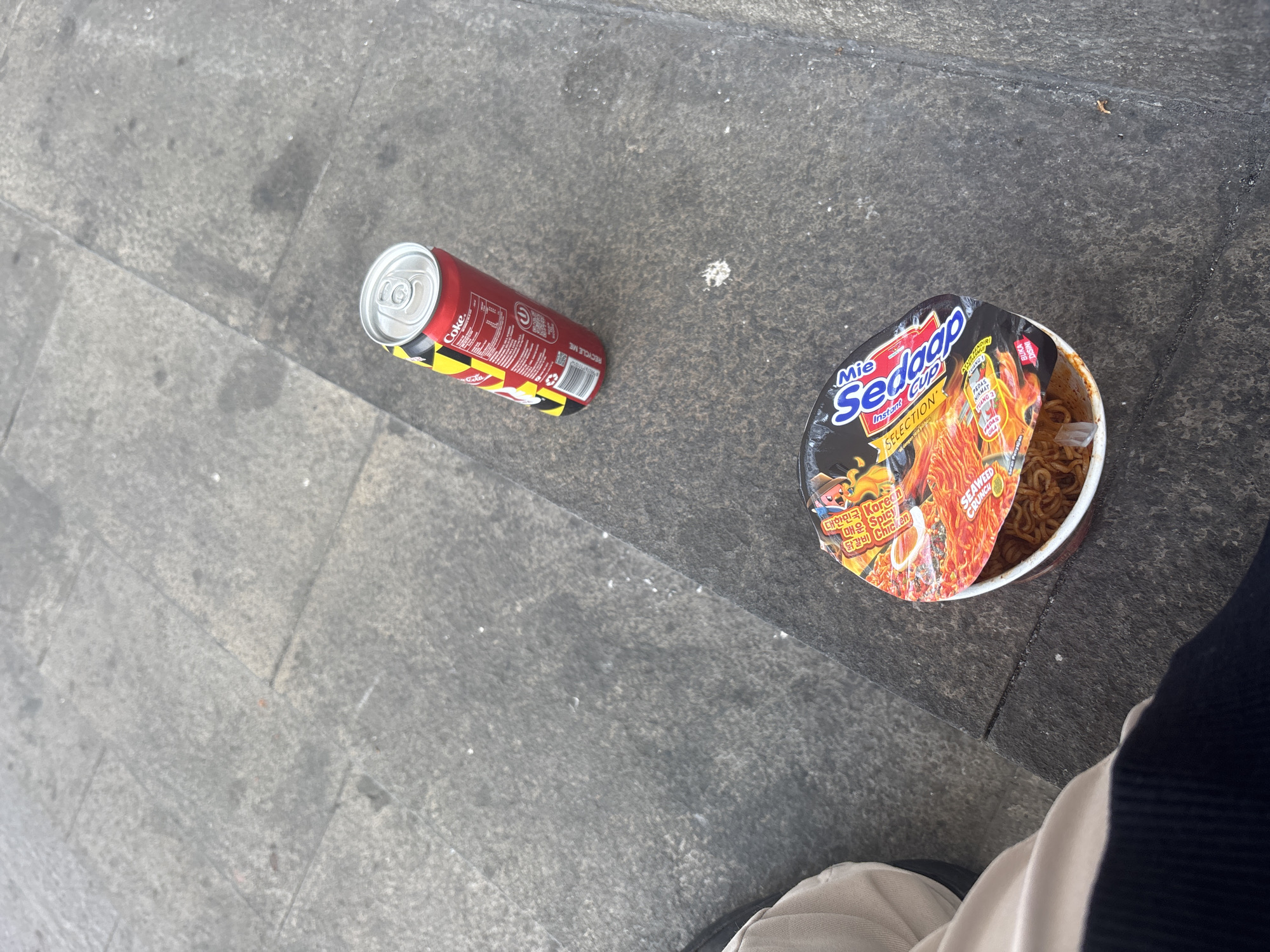 In the city of North Jakarta, Indonesia, an overhead shot captures a scene on a textured, grey outdoor surface, possibly a sidewalk. To the right, a red Coca-Cola can stands upright, its yellow and black warning stripes visible. To the left, a nearly consumed cup of instant noodles, labeled "Mie Sedaap SELECTION," rests on the surface. The lid of the noodle cup is partially open, revealing the contents. The bottom left of the frame shows the lower portion of a person's legs, clad in beige trousers and dark fabric, suggesting they are seated or have just finished eating. The overall environment appears to be a public space, with the lighting and textures suggesting an urban setting.