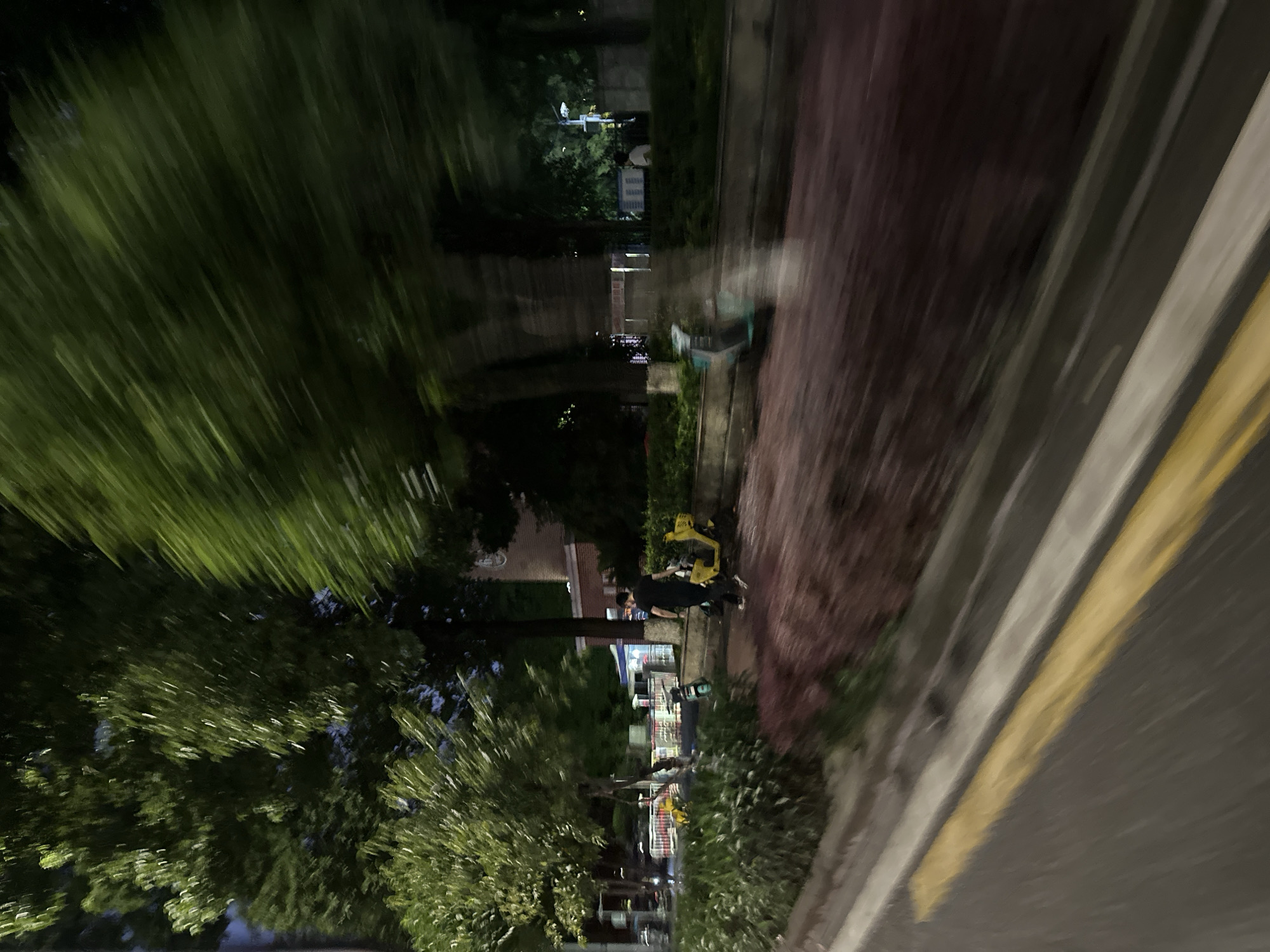 The image depicts an outdoor scene at dusk or night, with motion blur indicating movement. In the foreground, a road with a yellow line is visible, suggesting a street or pathway. Beyond the road, a sidewalk or pedestrian area is lined with lush green bushes. Further back, several parked electric scooters are present, two of which are yellow. A person, wearing a black t-shirt and pants, is walking away from the viewer towards the parked scooters. In the background, trees with green foliage dominate the scene, with buildings and streetlights faintly visible. The overall impression is that of a street in a well-lit urban environment, possibly a park or a residential area, given the presence of greenery and pedestrian infrastructure. The location is identified as the Chongqing High-tech Zone, China, which is an area known for its technological development and modern infrastructure, often featuring green spaces and accessible public transportation like electric scooters.
