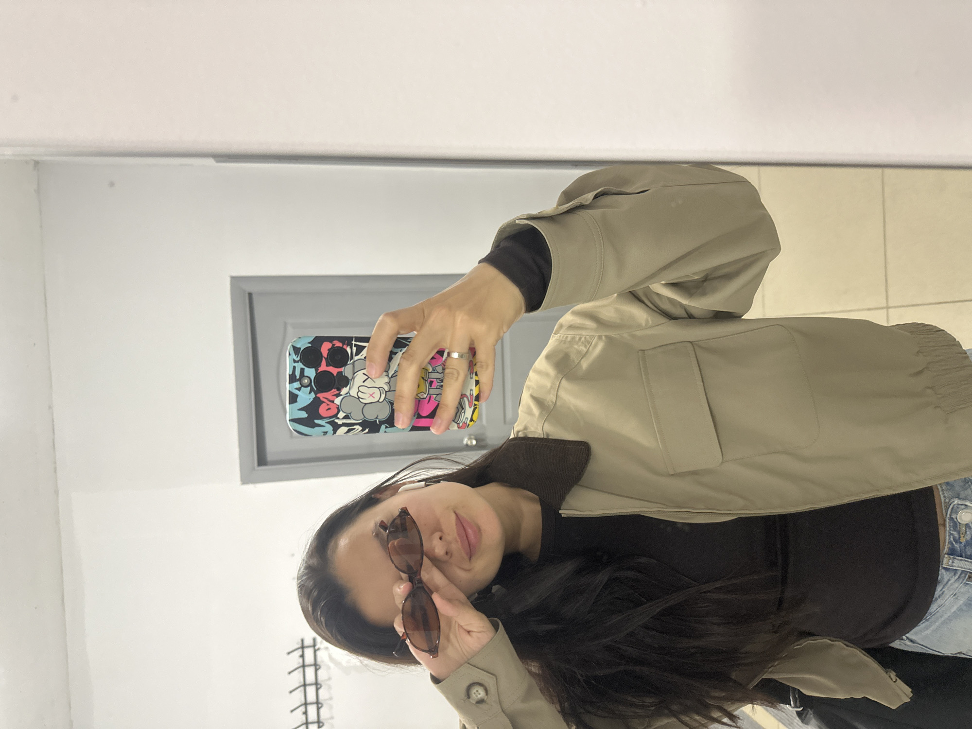 The image, rotated 90 degrees clockwise, depicts a woman taking a mirror selfie in an indoor setting. She has long, dark hair and is dressed in a light beige or olive-green jacket with a brown collar, layered over a black long-sleeved top and light blue denim jeans. Her left hand, wearing a silver-colored ring on the ring finger, holds a smartphone with a vibrant, graphic phone case featuring cartoon-like characters, which is visible in the mirror's reflection. With her right hand, she is pulling down a pair of dark, brown-framed sunglasses to look over them, displaying a subtle, relaxed expression.
The background consists of plain white or light grey walls and a light-colored tiled floor. A grey-framed door or structure is partially visible behind her phone, adding to the impression of an interior space such as a dressing room or private area. The activity is a casual self-portrait. No discernible text is visible within the frame. While the location context is provided as Chaikovsky, Russia, there are no specific visual cues within the image itself to confirm this detail. The overall impression is one of casual style and an unposed moment.