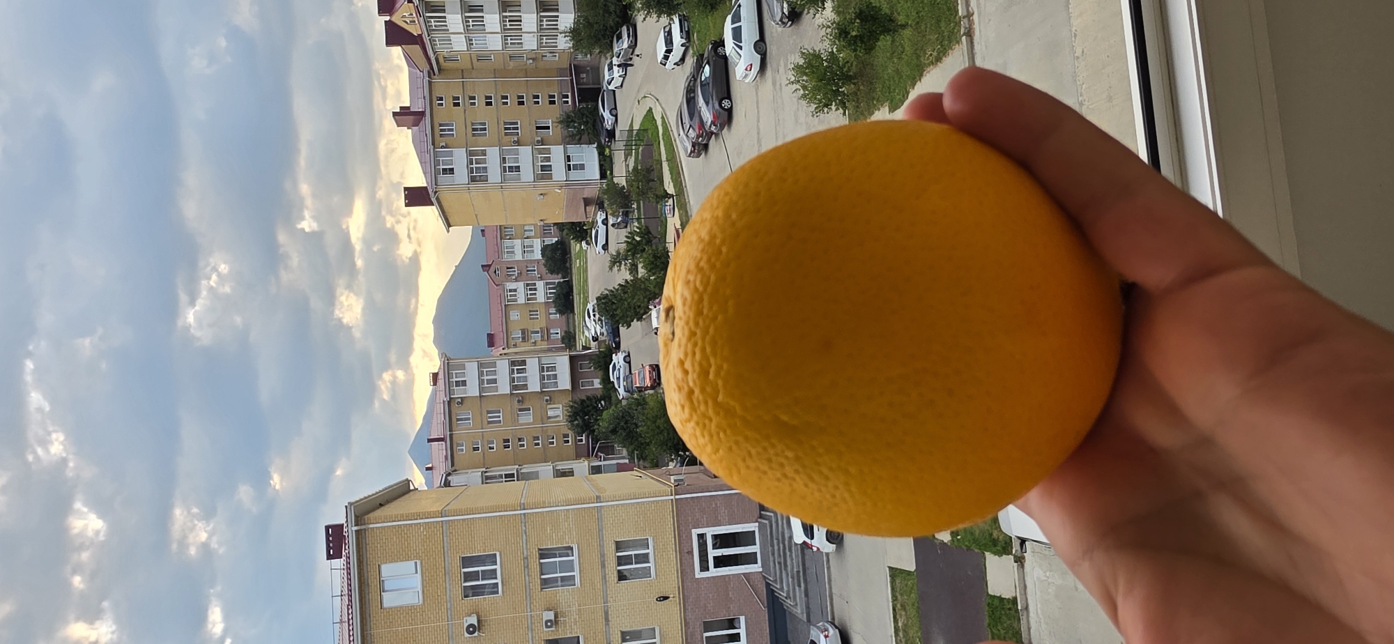 A hand holds a bright orange in the foreground, obscuring much of the background. The orange is textured and round. Behind it, a street scene unfolds in a residential area. Several multi-story apartment buildings, primarily yellow with red roofs, line a paved road. Numerous cars are parked on the street and in a lot. Green trees and bushes are interspersed throughout the scene. In the distance, a large, rounded mountain is visible under a cloudy sky with patches of sunlight breaking through. The location is Inozemtsevo, Russia, characterized by its apartment buildings, parked cars, and mountainous backdrop.