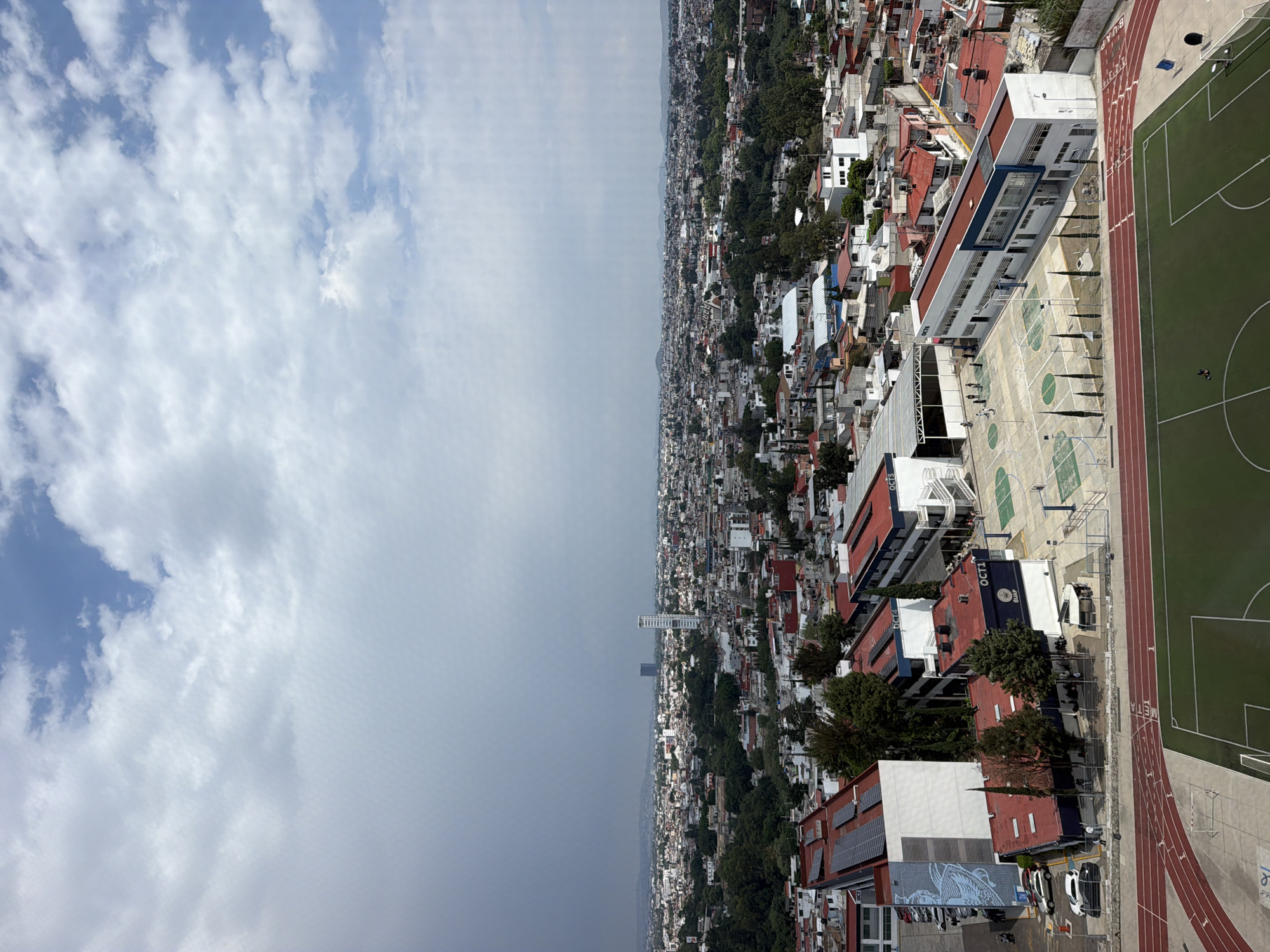 The image shows an aerial view of a densely populated urban area, likely the city of San Bernardino Tlaxcalancingo, Mexico. The left side of the image is predominantly sky with scattered clouds. The right side shows a built environment consisting of closely packed buildings, mostly residential, with red tile roofs. A tall, slender building is visible in the distance. A school campus is clearly visible, featuring a soccer field, a running track, and outdoor basketball courts. Several individuals appear to be present on the basketball courts.