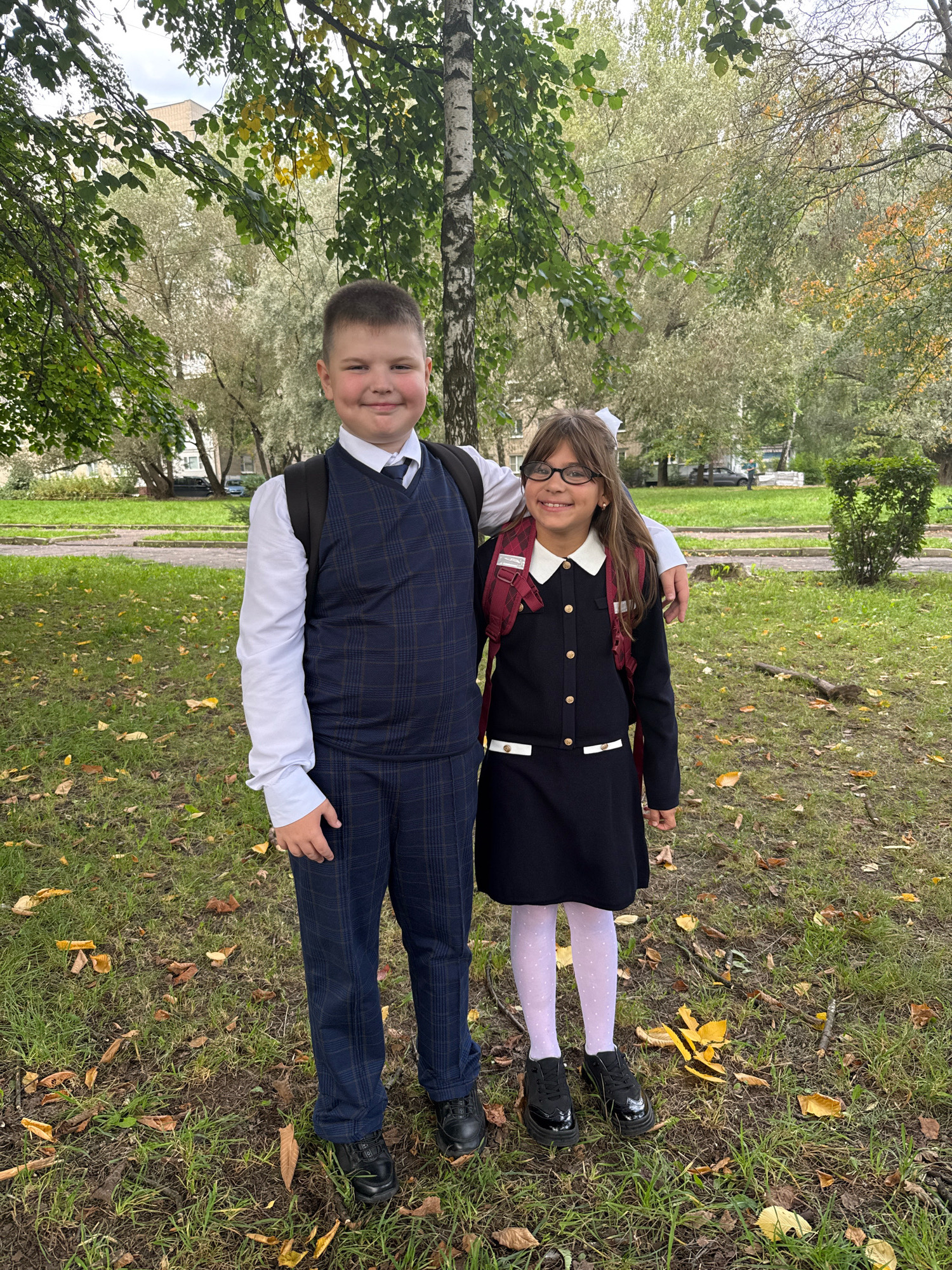 Two children, a boy and a girl, stand side-by-side in a grassy park area with fallen leaves scattered on the ground. The boy, positioned on the left, is wearing a white dress shirt, a dark plaid vest, and dark plaid pants. He has a dark backpack slung over his shoulders. The girl, on the right, is wearing a dark dress with a white collar and white trim at the hem, white polka dot tights, and black shoes. She also has a dark backpack, worn over her shoulders. She is wearing glasses and has a white bow in her hair. The boy has his arm around the girl's shoulders. In the background, there are trees with green and yellowing leaves, some buildings, and a paved path. The environment suggests an outdoor setting in a park or schoolyard during autumn, likely in or around Rusanovo, Russia, given the context.