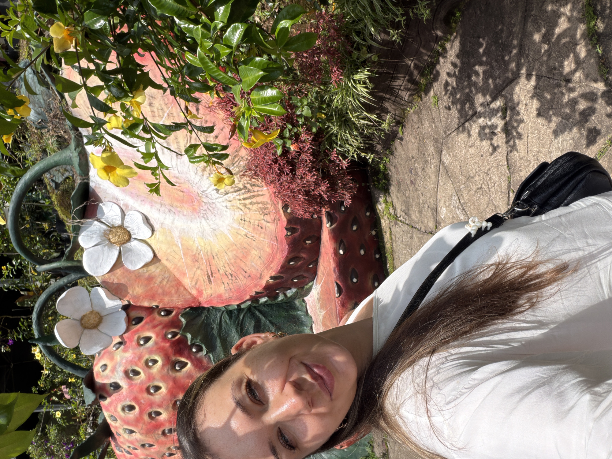 A woman with long brown hair and a white shirt smiles at the camera, taking a selfie. She is positioned in the foreground, with a large, colorful strawberry sculpture behind her. The sculpture appears to be cut in half, revealing the inner flesh and seeds. White flowers with yellow centers are attached to the sculpture. Lush green foliage and yellow flowers are visible to the right of the sculpture, casting shadows on a stone pathway in front of it. The scene is outdoors, likely in a garden or park, and the lighting suggests it is daytime. The presence of the strawberry sculpture and the vibrant greenery points to a tourist attraction, possibly in Dalat, Vietnam, known for its flower gardens and strawberry farms.