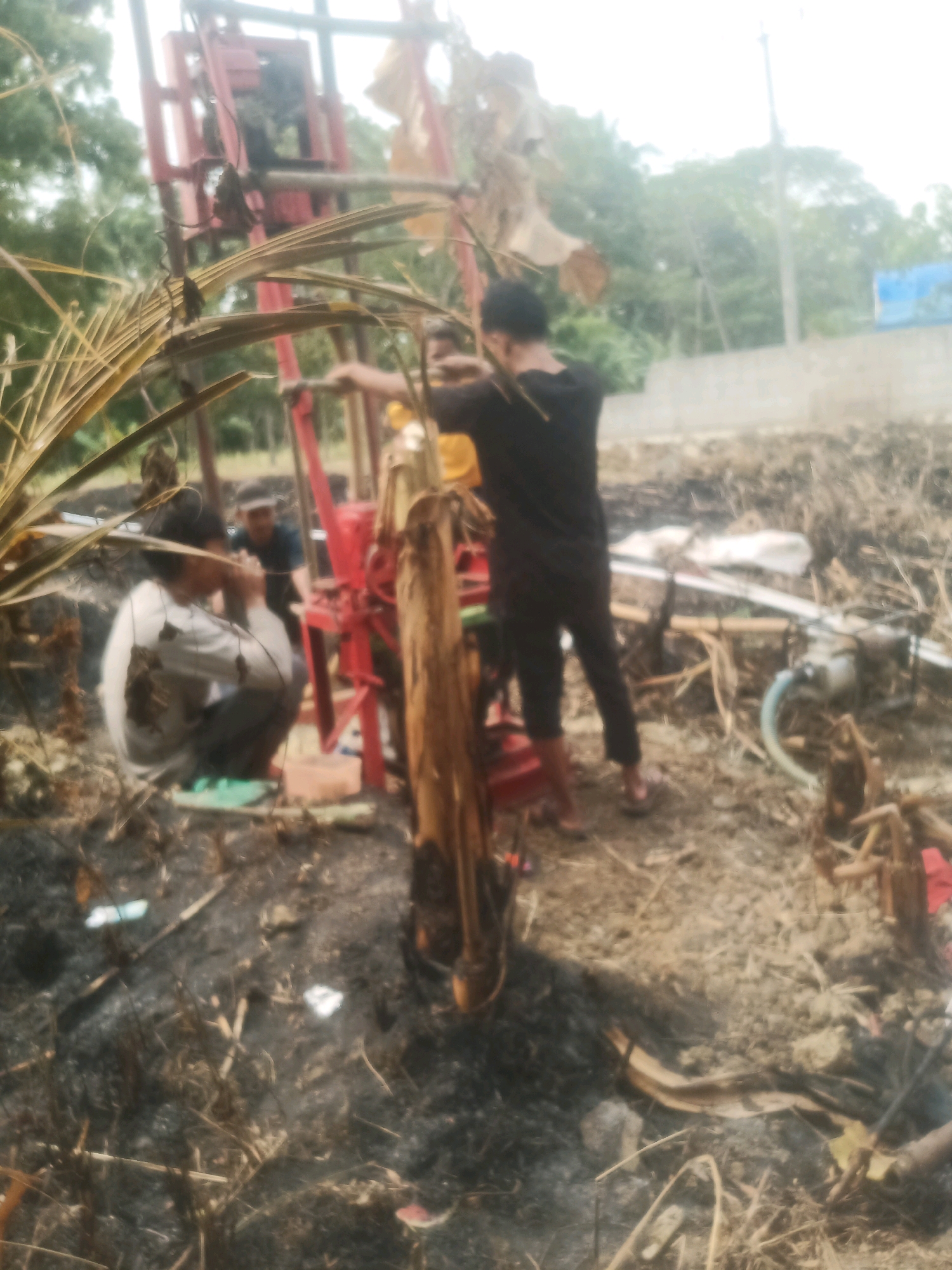 In Banyuasih, Indonesia, a group of individuals is engaged in drilling operations. The scene is characterized by dry, cleared ground with visible debris and burnt vegetation. A red drilling rig dominates the center of the frame, with a person in a black t-shirt and dark pants operating it. To the left, a person in a light-colored shirt and grey pants is crouched down, seemingly working near the base of the rig. Another individual in a dark t-shirt and a white cap is visible behind the rig. The environment suggests an outdoor, possibly rural or semi-developed area, with trees in the background and a light-colored structure or wall further in the distance. There is also evidence of water pipes and a pump-like device on the ground.