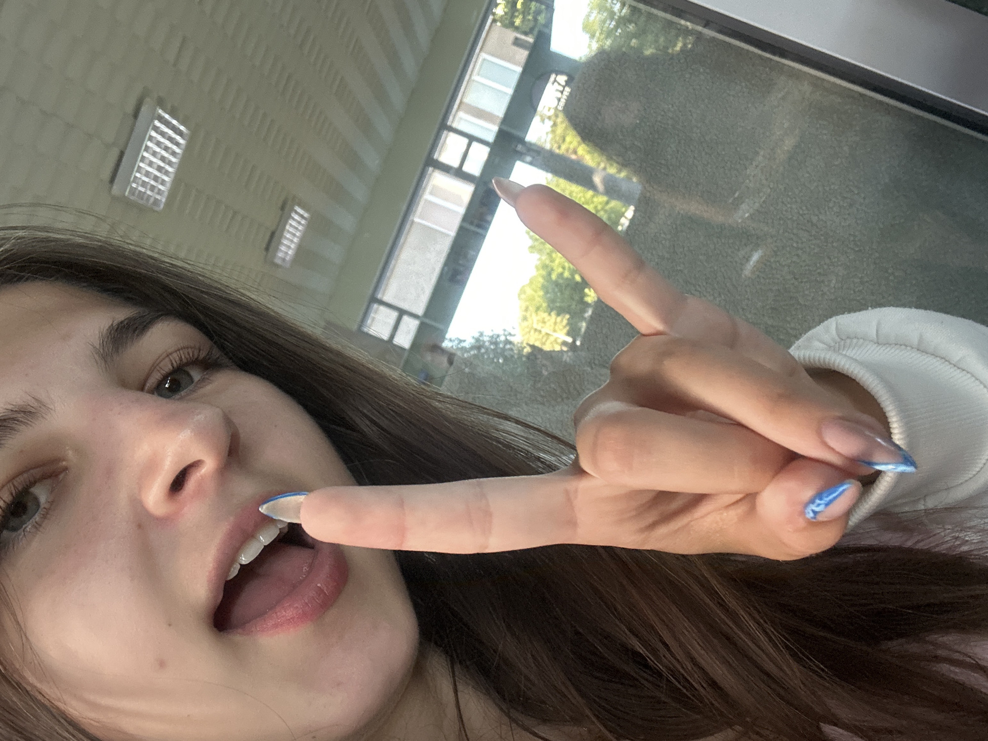 In this close-up, indoor selfie, a young woman with long brown hair extends her right hand, fingers forming a "rock on" gesture. Her left index finger is touching her lips, which are slightly parted, revealing her teeth and tongue. She has blue and white patterned artificial nails on her visible fingers. The background reveals a tiled ceiling with fluorescent light fixtures and a large glass window or door, through which trees and a building facade are visible. The environment suggests an indoor public space, possibly a cafe or a shop, given the reflection of a sign with the word "COFFEE" on the glass. The lighting is bright and diffused. The image does not contain any elements that would suggest a specific location within Shumen, Bulgaria.