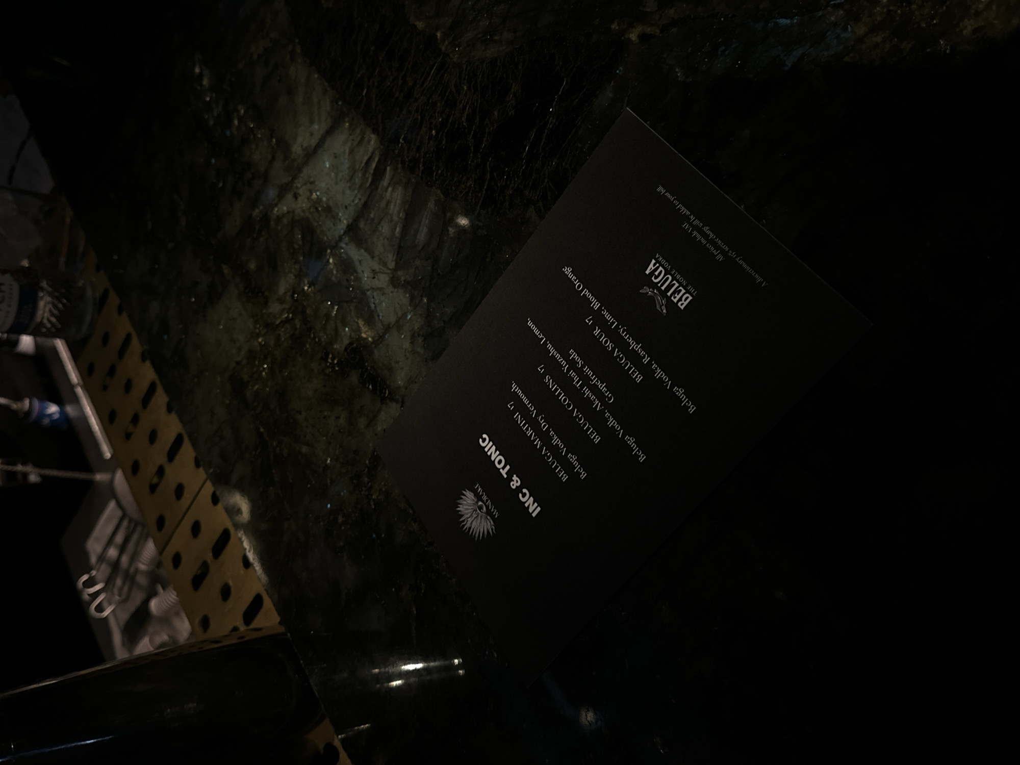 The image shows a black menu card for Mandrake, a bar in London, United Kingdom. The menu advertises Beluga vodka drinks. The card is placed on a dark, reflective surface that could be a table. The background is dark and out of focus, but there is a hint of a structure or object to the left. The lighting is low, creating shadows.