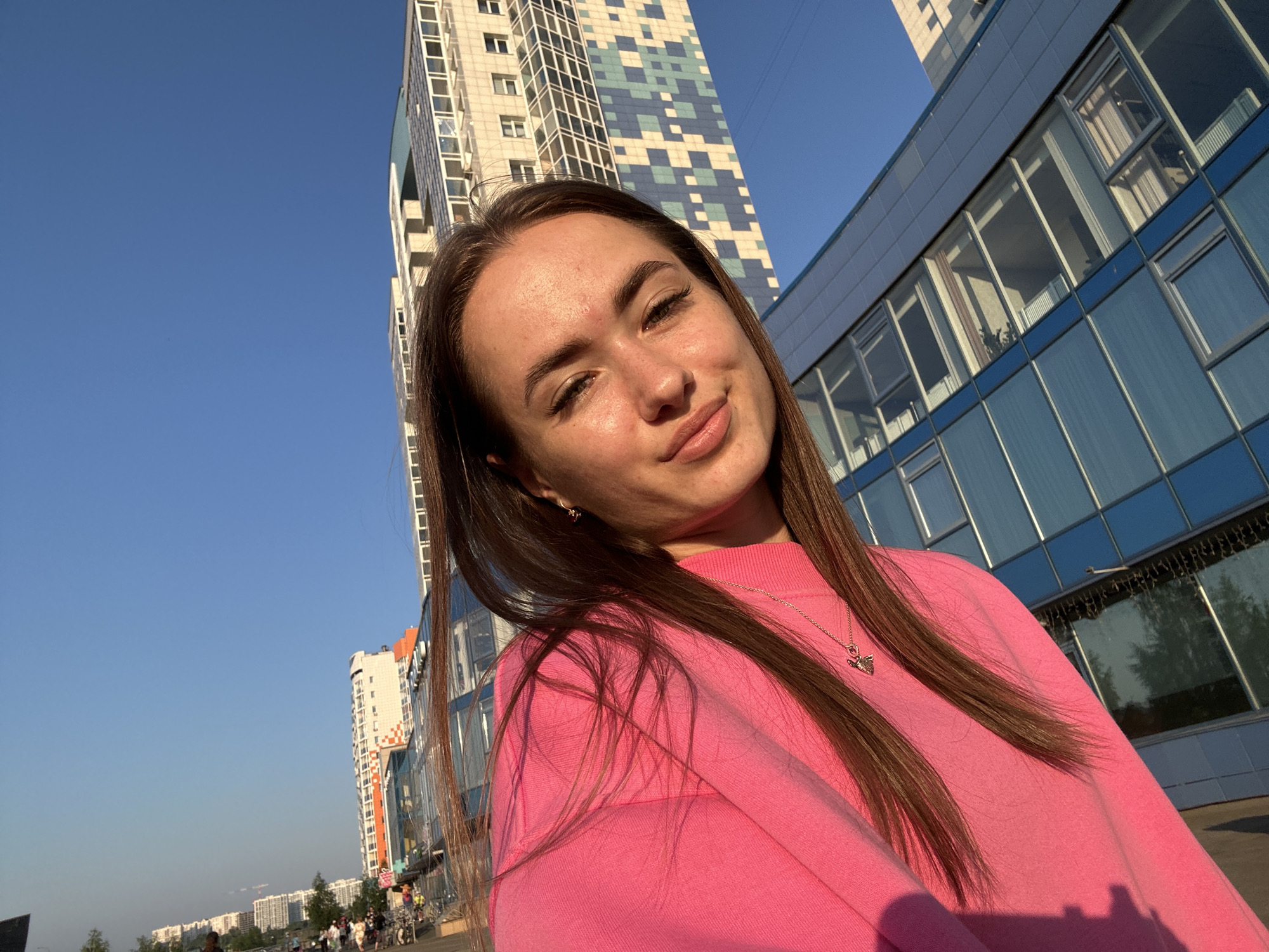 A young woman with long, brown hair and light skin is taking a selfie outdoors during the day. She is wearing a bright pink long-sleeved shirt and a silver necklace. Her head is tilted slightly, and she is smiling with her lips pursed. In the background, there is a modern, tall building with a colorful, geometric facade, and another building with a blue glass exterior. The sky is clear and blue. The scene appears to be in an urban area, possibly a city.
