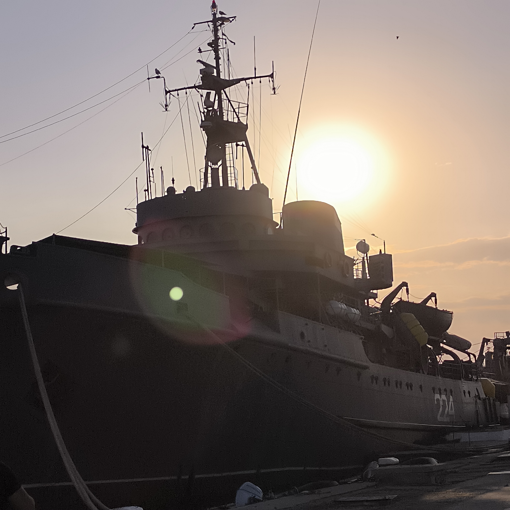 This is a photograph of a large, dark-colored ship docked at a pier, likely in Varna, Bulgaria, given the context. The sun is setting or rising, casting a warm, orange glow across the sky and creating a silhouette of the ship. The ship appears to be a naval vessel, with a prominent mast structure, antennas, and what seem to be lifeboats or rafts attached to its side. The number "224" is visible on the hull of the ship. The sky is mostly clear with some faint clouds, and a bird is visible flying high in the distance. The foreground shows a portion of the pier, made of concrete and wood, with some ropes and other maritime equipment. The overall impression is one of a tranquil maritime scene at dawn or dusk.