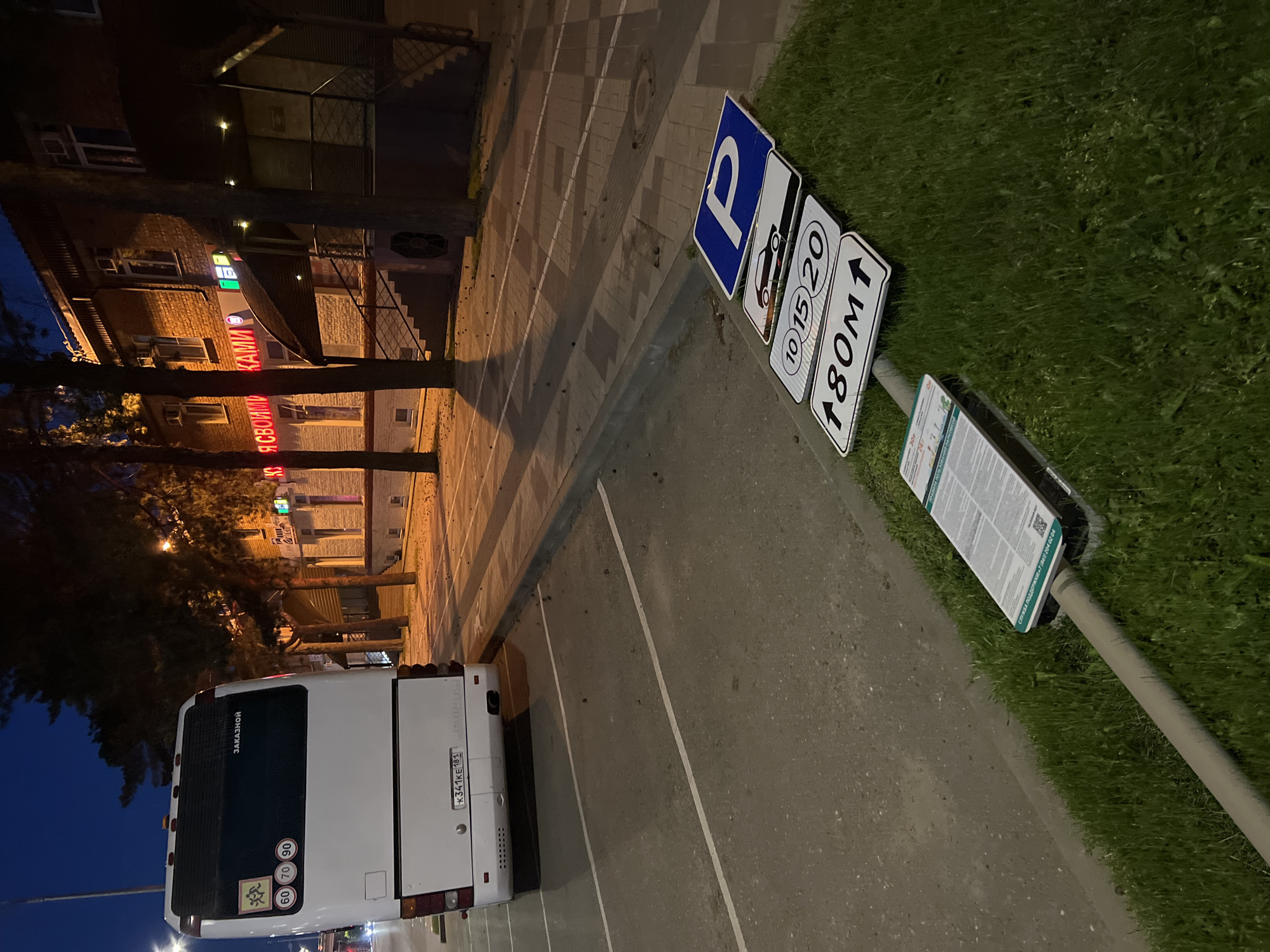 The image captures a parking area scene at night. A white bus with Russian license plate number K341KE 181 is parked in one of the parking slots. The bus has a "ZAKAZNOY" sign, indicating it is a chartered bus. Speed limit stickers of 60, 70, and 90 are visible on the bus. A parking sign with the letter "P" in blue is displayed. Another sign indicates parking is restricted between 10:00 AM and 8:00 PM for the next 180 meters. A third sign provides service support information. The image is taken at an angle, revealing part of a building that reads "Я СВОИМИ КАМИ" in red neon letters, which translates to "With Your Own Hands". The location is Krasnodar, Russia. The environment includes grass patches and a paved walkway with brickwork.