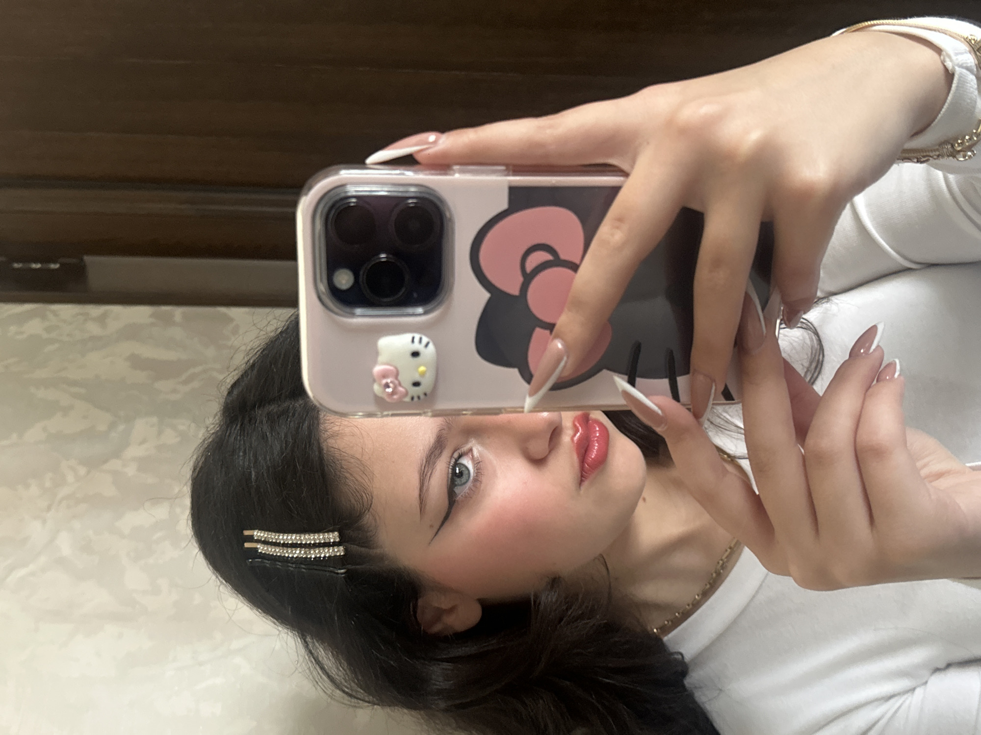 The image depicts a woman with dark hair and blue eyes taking a selfie with her phone. The phone case is pink with a Hello Kitty design. She has fair skin, prominent cheekbones, and red lipstick. Her hair is styled with a jeweled clip. She is wearing a white top. The background appears to be a room with a patterned wall and a dark wood-toned piece of furniture or wall paneling. There is no information to tell us whether this image was taken in Yerevan, Armenia.