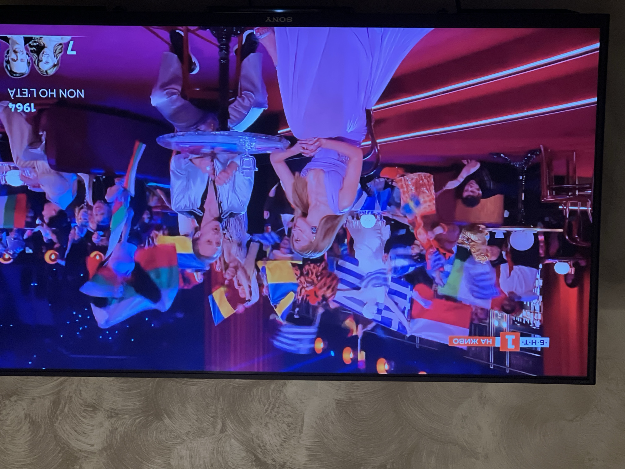 The image depicts a television screen displaying what appears to be a broadcast of the Eurovision Song Contest. The broadcast features performers on stage, an audience with flags, and a background with stage lighting. The text "БНТ 1 НА ЖИВО" is visible on the lower right of the screen, confirming it is Bulgarian National Television. The text "NON HO L'ETA' 1964" is visible on the upper left of the screen. The location of the event is likely not Sofia, Bulgaria, as the Eurovision Song Contest is typically held in a different European city each year. The image includes details like performers in costumes, audience members holding flags of various nations, and elements of stage design.