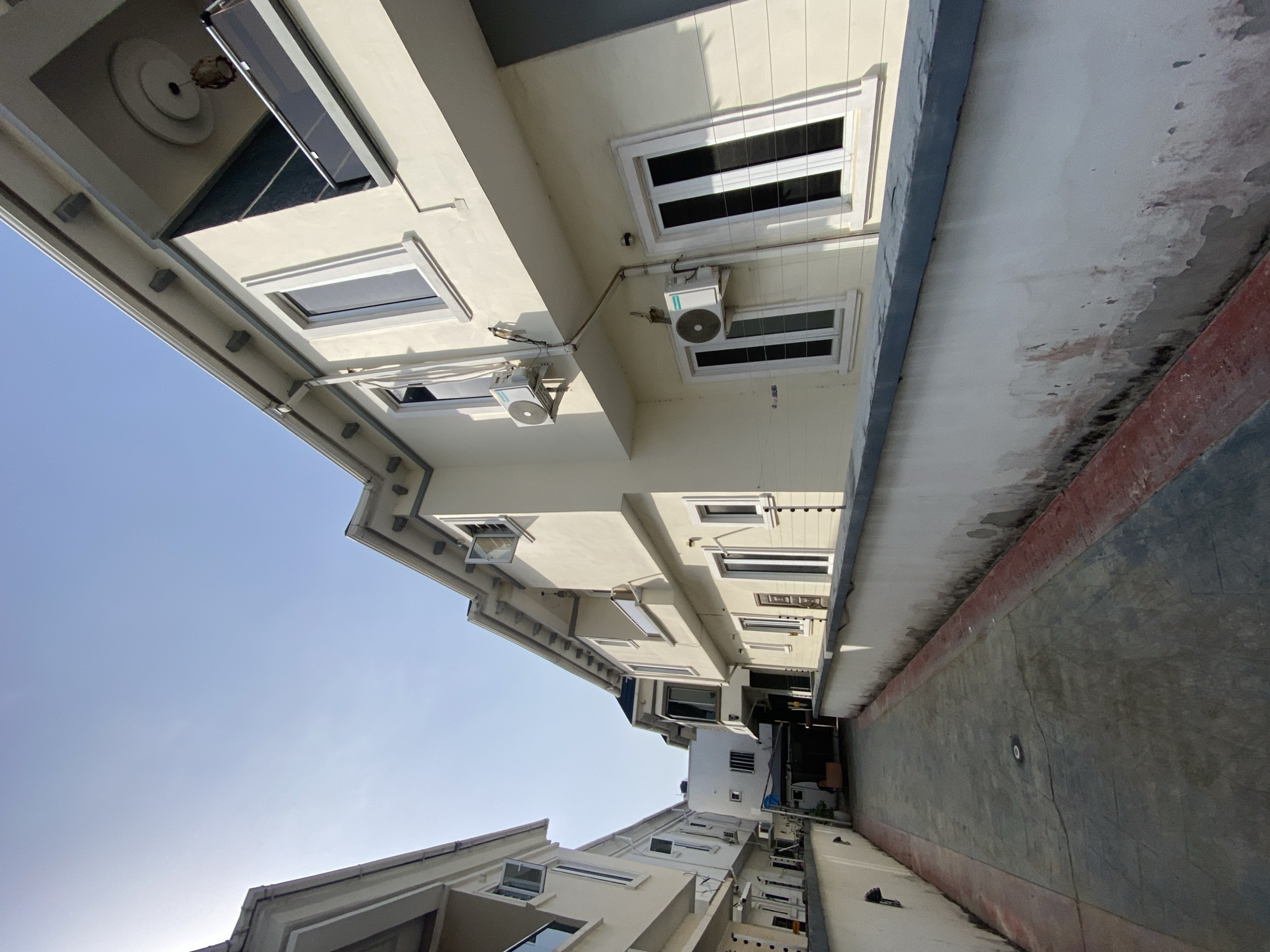 The image shows a narrow alleyway between buildings, possibly in Lagos, Nigeria. The buildings are light-colored with windows, some open. Air conditioning units are visible on one of the buildings. The alleyway floor is concrete with a dark border along the building. The sky is light blue. No people are visible.