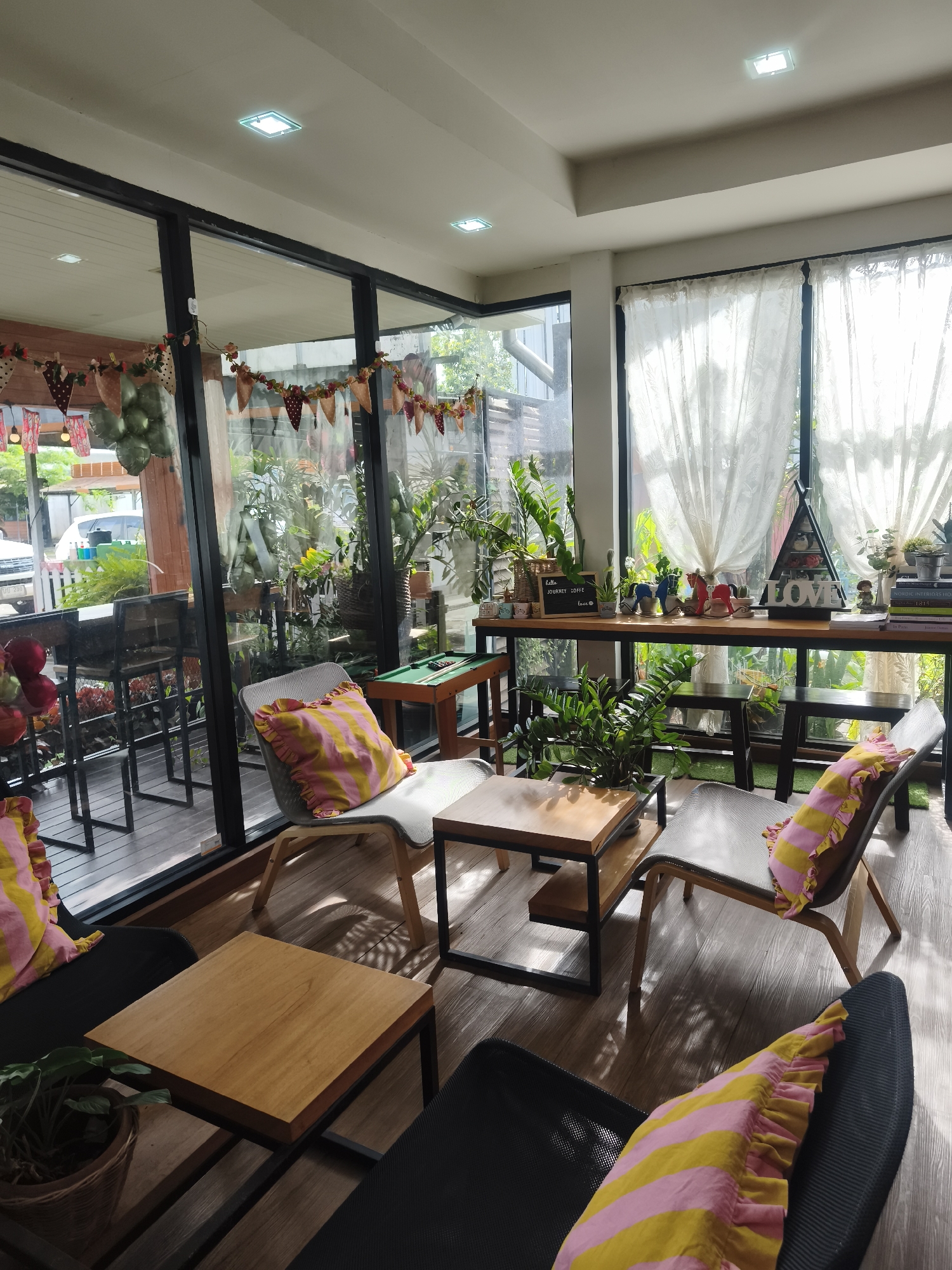The image depicts the interior of a modern cafe in Mai Khao, Thailand, likely an establishment designed to be a comfortable and inviting space for patrons. The scene is well-lit, with natural light streaming in through large windows. The decor suggests a contemporary aesthetic with natural elements, possibly aiming for a relaxed ambiance.
The cafe features a seating area with several chairs and tables. Two chairs with grey upholstery are prominently placed in the foreground, each adorned with a vibrant yellow and pink striped cushion. These chairs are positioned around a small, square wooden coffee table with a black metal frame. Another similar coffee table is visible to the left, near a dark-colored chair with another striped cushion.
Beyond the immediate seating area, a long wooden counter runs along the back wall, acting as a bar or display area. On this counter, various decorative items are arranged, including potted plants, figurines, and what appears to be a "LOVE" sign. Behind the counter, more seating, possibly bar stools, is visible, suggesting a separate dining or drinking area.
The cafe's design incorporates extensive glazing, with large windows that offer views of an outdoor space with lush greenery and some trees. This suggests the cafe might be situated in a tropical or garden-like setting, which is common in many parts of Thailand. The floor is wooden, contributing to the warm and natural feel of the space. Decorative garlands with small triangular pennants are strung along the top of the windows, adding a festive touch.
The overall impression is one of a well-maintained and aesthetically pleasing cafe, likely designed to attract customers seeking a comfortable and stylish environment for relaxation or socializing. No people are visible in the image, focusing the attention solely on the cafe's environment and furnishings.