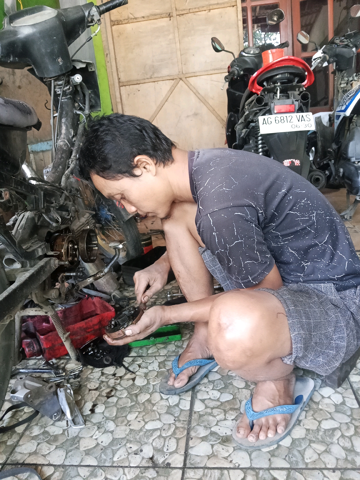 In Wates, Indonesia, a man is seen working on a motorcycle in what appears to be an outdoor workshop. He is kneeling on the ground, focused on the engine of a disassembled motorcycle. Various motorcycle parts, tools, and red plastic containers are scattered on the ground around him. The ground is paved with light-colored, irregular stones. In the background, other motorcycles are visible, and a wooden wall with panels forms the backdrop. The man is wearing a dark t-shirt with a white speckled pattern and grey checkered shorts, along with blue flip-flops. The scene suggests a routine repair or maintenance activity taking place in a typical Indonesian setting.