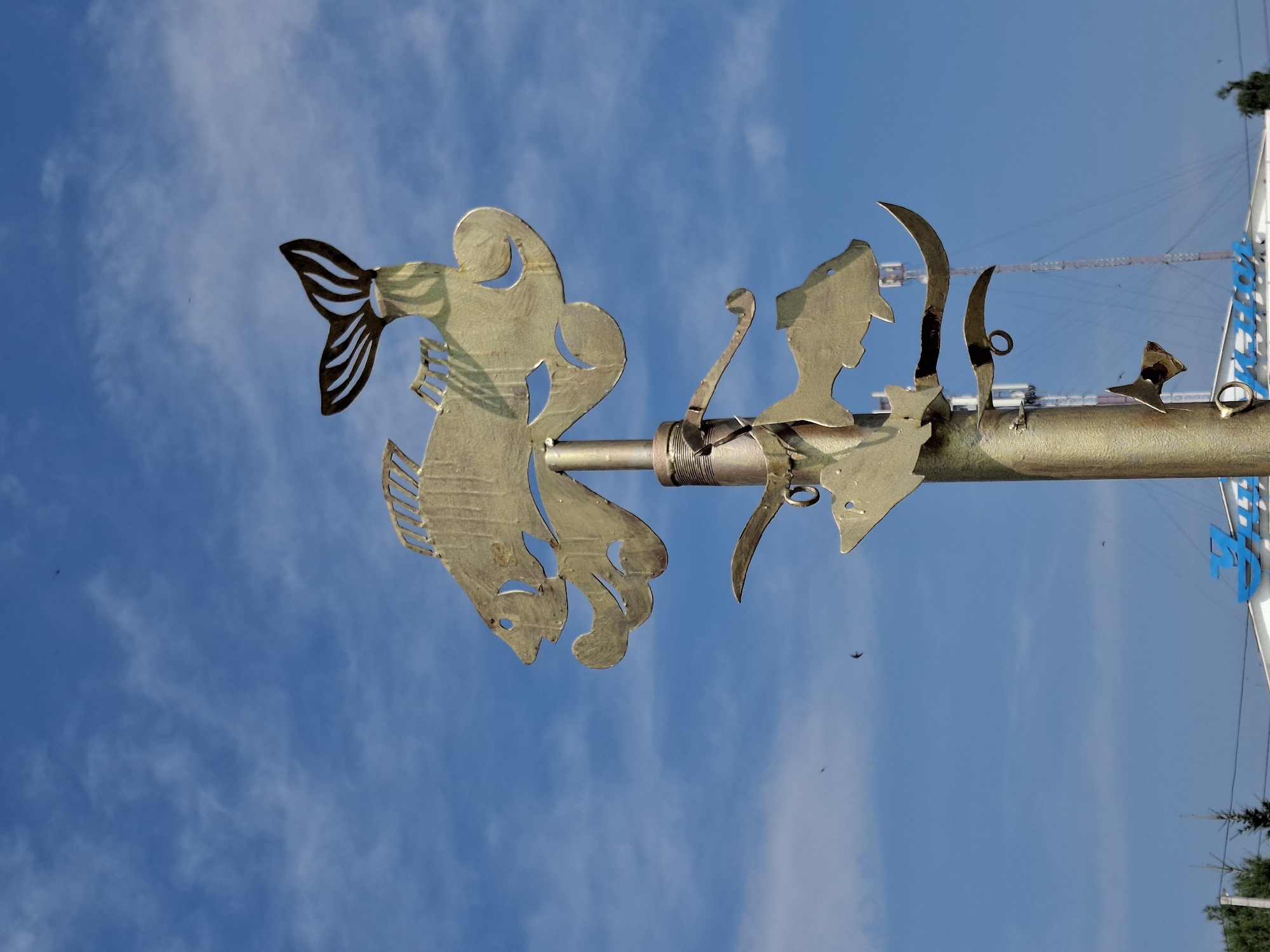 A metal sculpture, resembling a school of fish, is mounted on a pole against a bright blue sky with scattered white clouds. The sculpture is made of golden-colored metal and features stylized fish shapes of various sizes. The largest fish is at the top, with its tail fin prominent. Below it, two smaller fish appear to be swimming, one with a curved body and the other more elongated. The pole itself is cylindrical and weathered, with visible bolts and connections. In the background, a distant cell tower and some trees are visible. Below the sculpture, a portion of a building with blue lettering can be seen. The image is a low-angle shot, emphasizing the sculpture against the sky.