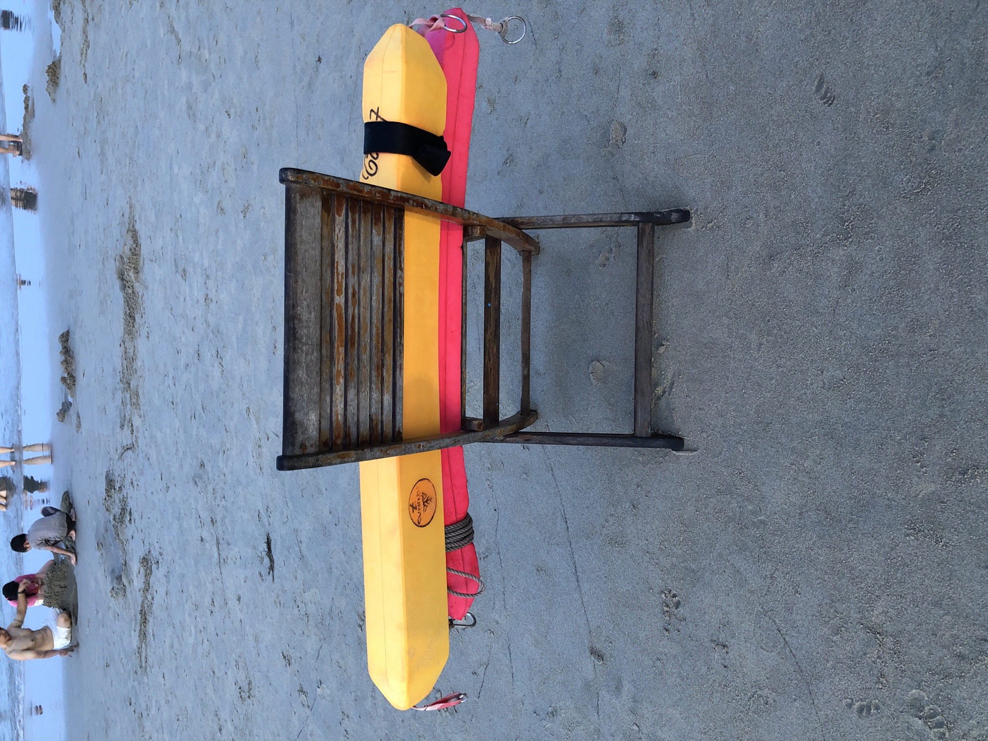 The image depicts a wooden lifeguard chair on a sandy beach, likely located in Da Nang, Vietnam, given the context. A yellow and orange flotation device, commonly used by lifeguards, is placed horizontally across the seat of the chair, secured by a black strap. A pink towel or blanket is also draped beneath the flotation device. In the background, shallow ocean water meets the shore, and several people, including children, can be seen playing in the sand and water. The overall atmosphere suggests a typical day at the beach.