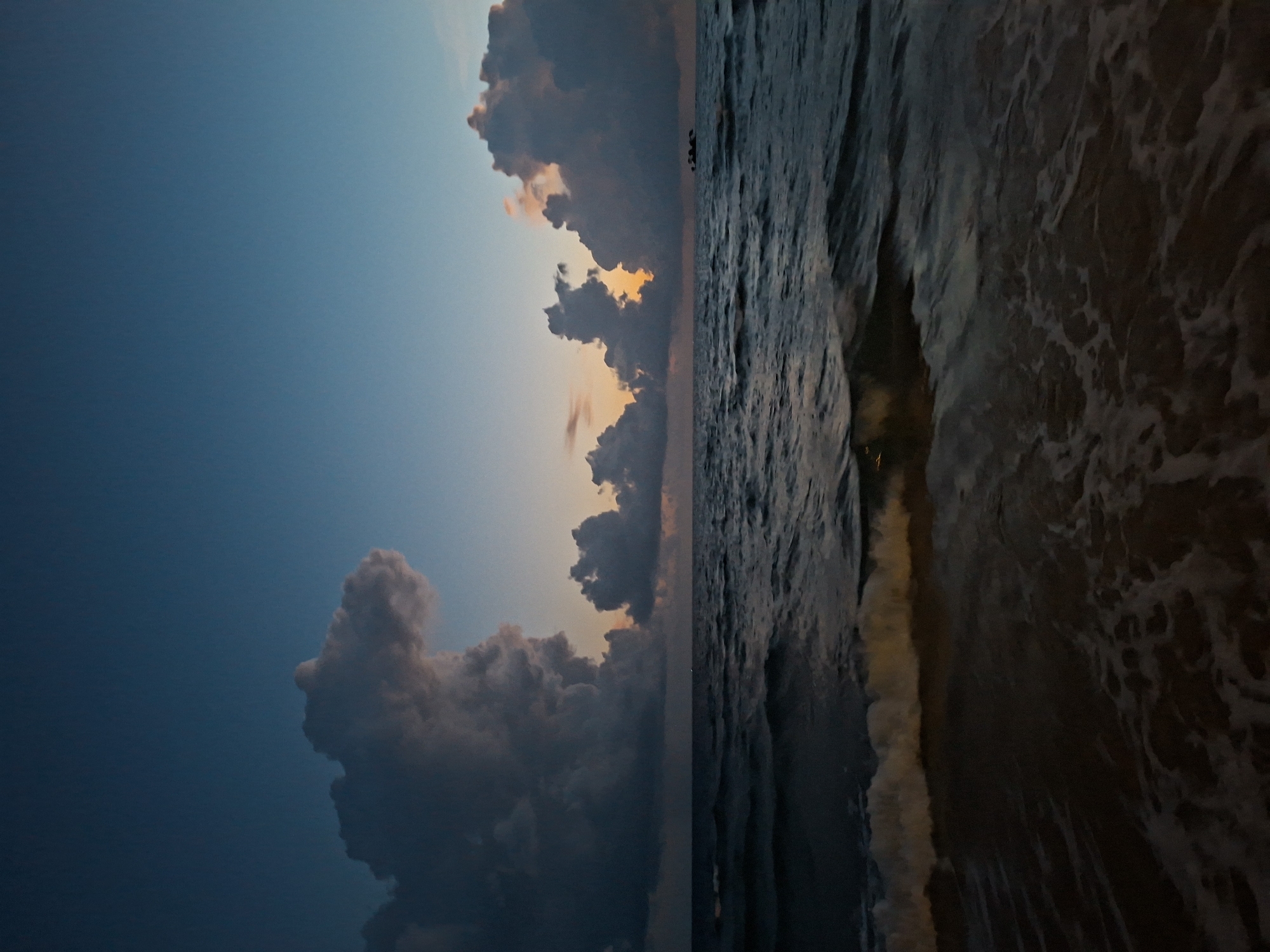 A wide shot captures a serene beach scene at dusk or dawn. The sky is a gradient of deep blue at the top, transitioning to lighter hues and hints of orange and yellow near the horizon, where dramatic, dark clouds are illuminated from behind. The sea is dark and choppy, with waves gently rolling towards the shore. White foam from the breaking waves is visible in the foreground, creating a soft, textured edge between the water and the darker sand. No people or structures are visible in the frame, emphasizing the natural beauty of the environment.