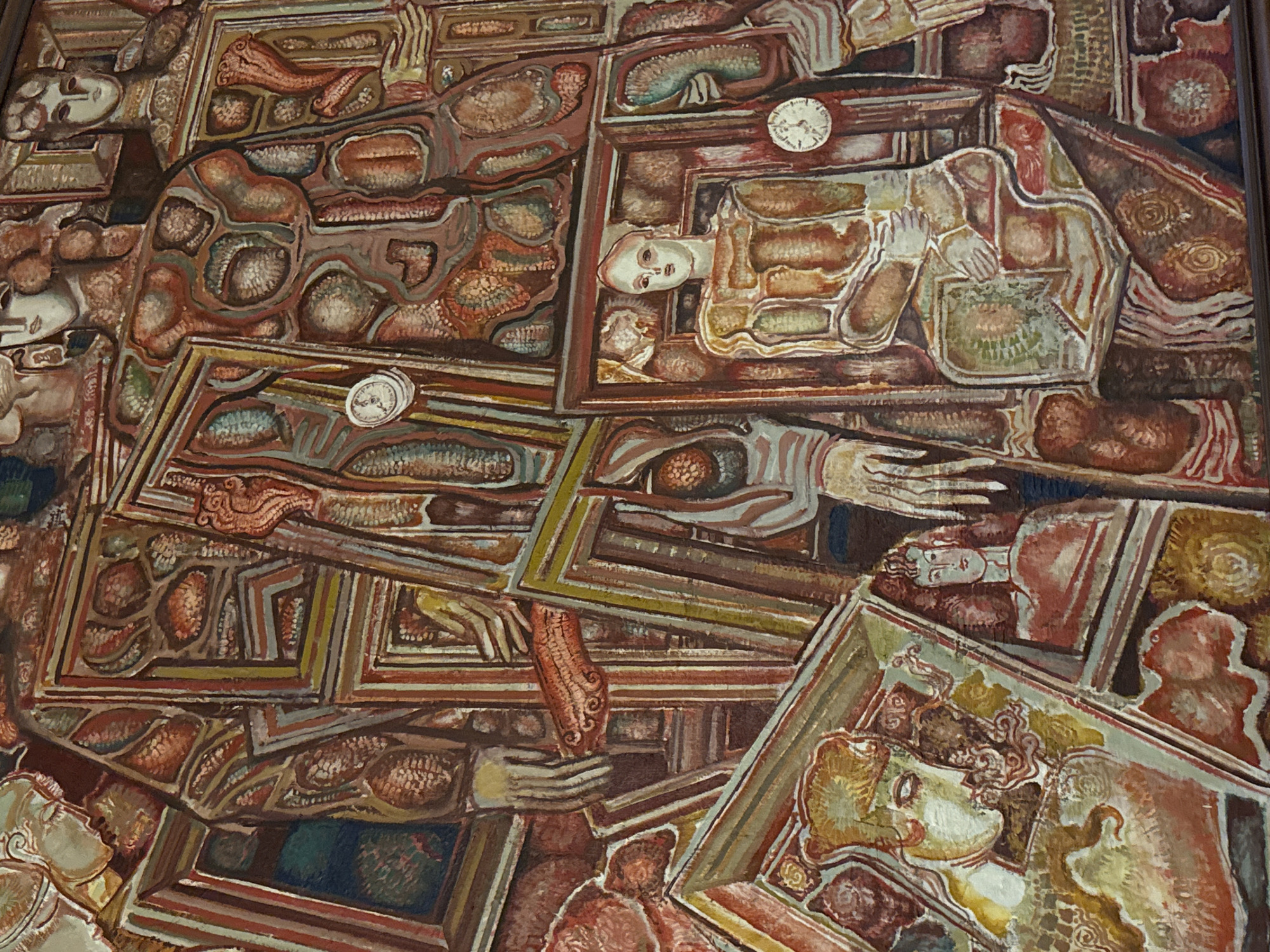 This intricate painting, possibly a mural or large canvas, presents a densely packed and surreal composition featuring numerous stylized human figures and fragmented body parts. The artwork is characterized by an earthy color palette of browns, reds, oranges, and muted greens and blues, with strong, graphic outlines defining its complex structure.
The central subjects are human figures, many depicted reclining or in a state of repose, reminiscent of sleep, dreams, or perhaps death. These figures are often enclosed within an elaborate network of interlocking, multi-layered frames, resembling drawers, coffins, or display cases. A prominent figure in the upper right quadrant features a pale, serene face, with a pocket watch positioned directly above their head. Another figure below and to the right is shown with hands clasped, possibly in prayer or contemplation. Throughout the artwork, other faces and disembodied hands emerge from or are integrated into the textured, patterned surfaces.
Notable objects include two visible pocket watches: one in the upper right and another in the middle left, both showing hands set to a similar time, suggesting a frozen moment or the cyclical nature of time. The entire composition is structured by these complex, overlapping frames that contain both the figures and abstract, organic shapes resembling biological forms or textured elements.
The painting does not depict a singular activity or event but rather evokes a contemplative, introspective state. The pervasive themes appear to relate to consciousness, memory, and mortality, conveyed through a fragmented, dream-like, and somewhat claustrophobic narrative. The overall mood is surreal and melancholic. No visible text is present within the artwork. This piece is located in Sofia, Bulgaria.