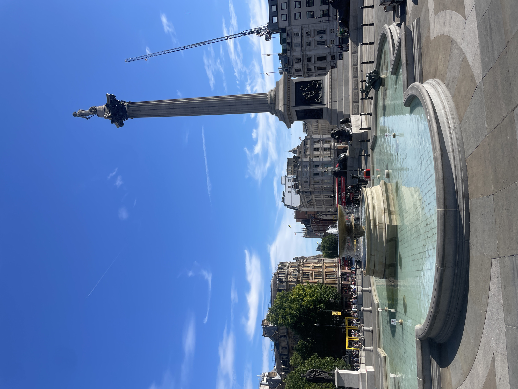 This media file depicts a sunny day at Trafalgar Square in London, United Kingdom. The central subject is Nelson's Column, a towering monument topped by a statue of Admiral Lord Nelson. The column is surrounded by a large fountain with clear, turquoise water, reflecting the bright blue sky and scattered white clouds. Bronze lion sculptures are positioned at the base of the column.
In the background, grand 19th-century buildings with neoclassical architecture line the square. People are visible, though they are small in scale due to the vastness of the location and the perspective. Some are gathered near the fountain, while others appear to be walking through the square. A red double-decker bus, a quintessential London sight, can be seen in the distance. The presence of a construction crane in the sky suggests ongoing development or maintenance work in the area.
The bright sunlight and clear skies indicate it is daytime, likely mid-morning to afternoon. The atmosphere appears lively and tourist-filled, typical of a popular landmark. The overall impression is one of a vibrant and iconic urban space on a pleasant day. No visible text is clearly legible in the image.