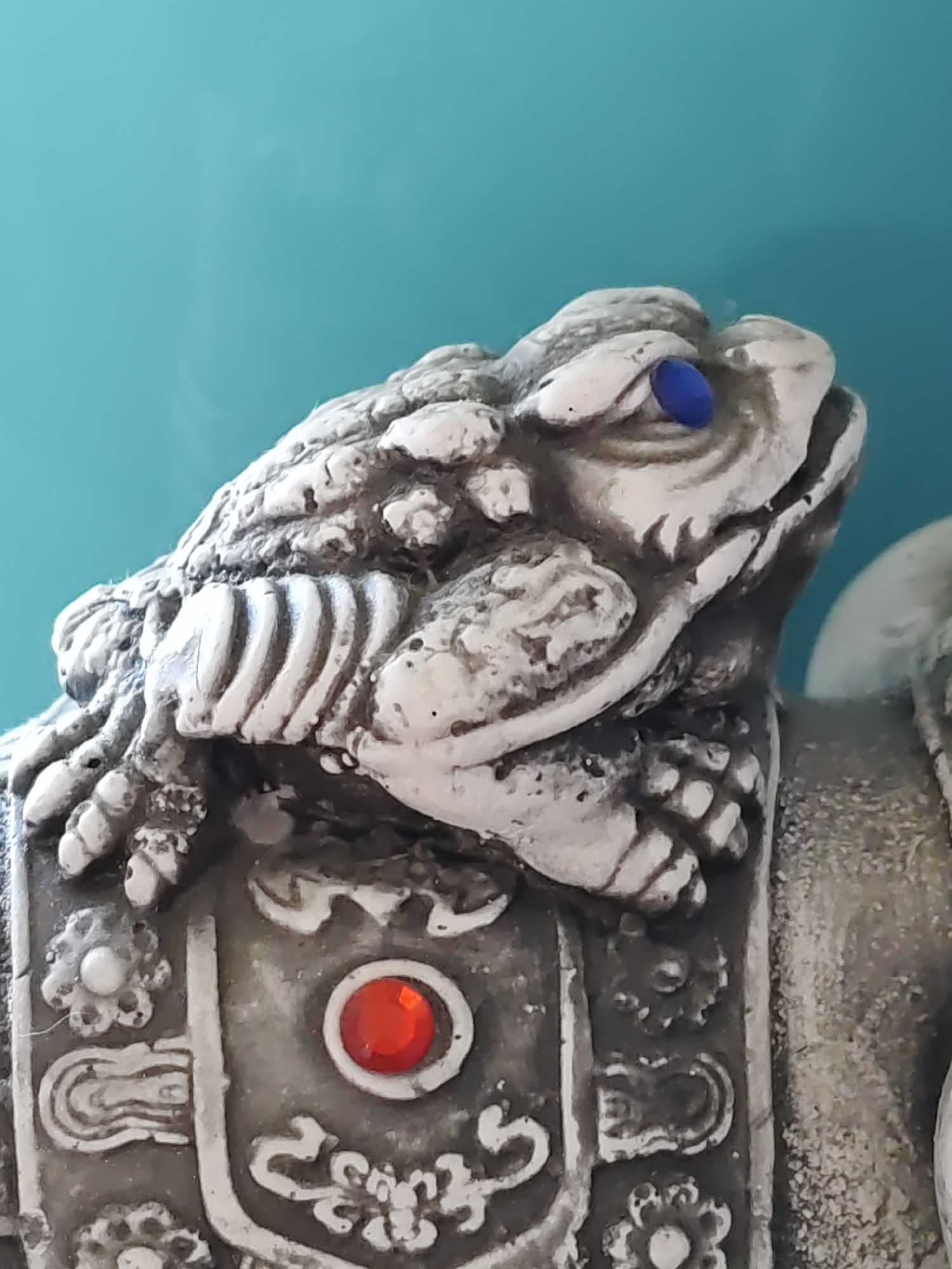 The image shows a close-up of a decorative statue of a frog, likely a mythical creature, with a blue bead for one eye and a red jewel embedded in its chest ornamentation. The statue appears to be made of stone or a similar material and features intricate carvings. The background is a solid, bright teal color, suggesting it is placed against a wall or fabric. The statue is positioned at an angle, with the frog's head turned to the right and its body facing forward. There is no discernible location in Rostov-on-Don, Russia.