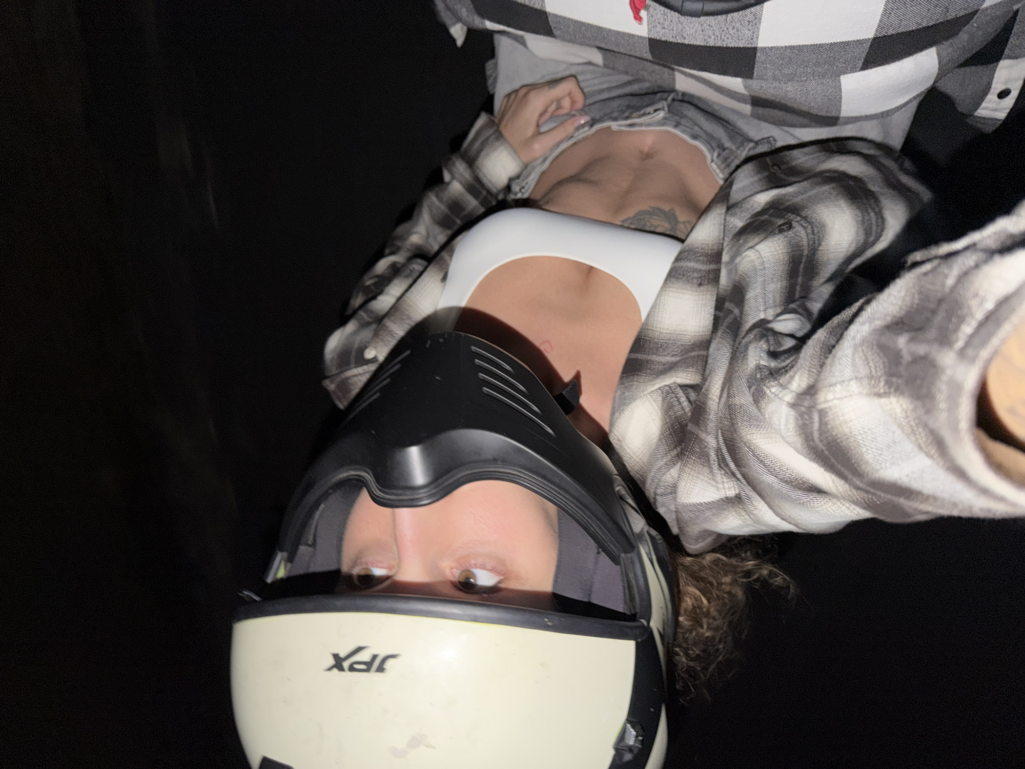 The image captures a close-up, sideways portrait of a person wearing motorcycle gear. The individual, appearing female, is wearing a light-colored, possibly off-white, full-face motorcycle helmet with the brand "JFX" prominently displayed on the front. Their brown eyes are visible above the helmet's chin bar, looking directly forward, with wisps of curly brown hair emerging from the back of the helmet.
The person is dressed in a white crop top or sports bra, over which a black, white, and grey plaid long-sleeved shirt is worn unbuttoned, revealing the midriff. A small red heart tattoo is visible on the chest, along with another tattoo design on the rib cage area. Light-colored pants or shorts are partially visible at the waist. One hand, featuring light-colored fingernails, is visible near the waistline.
The background is uniformly dark and out of focus, suggesting a night time setting or an extremely low-light environment. Given the provided location context of North Kuta, Indonesia, it likely depicts someone posing outdoors in the evening, possibly before or after a motorcycle ride. The person's expression appears neutral.