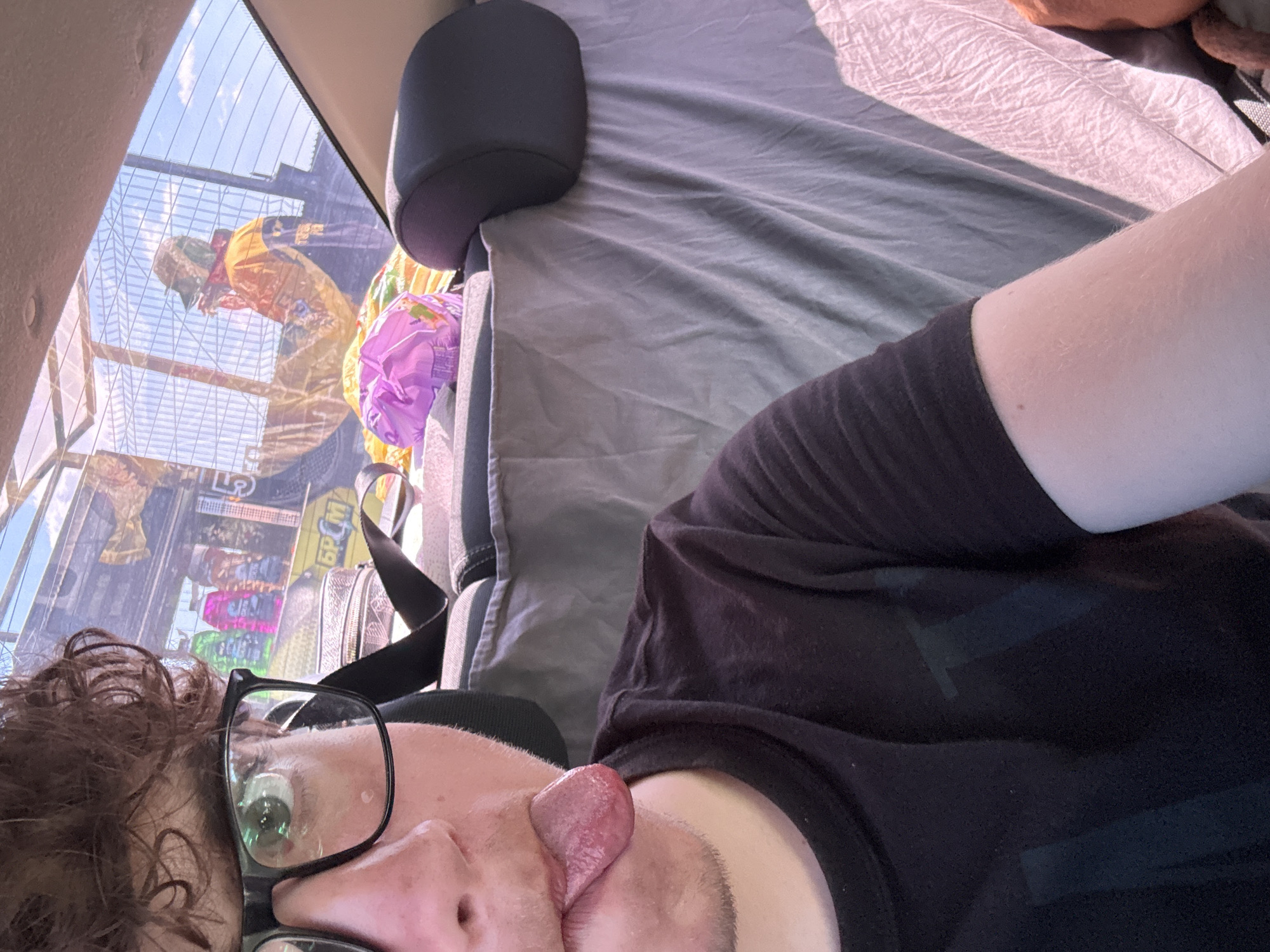 The image is a selfie taken from inside a vehicle. The foreground features a person with curly brown hair and glasses, sticking their tongue out. They are wearing a black t-shirt. In the background, visible through the rear window, is a busy street scene with buildings and a bright blue sky. The overall lighting suggests it is daytime. The exact location in the city of Velykodolynske, Ukraine, cannot be determined from the visual information present in the image.