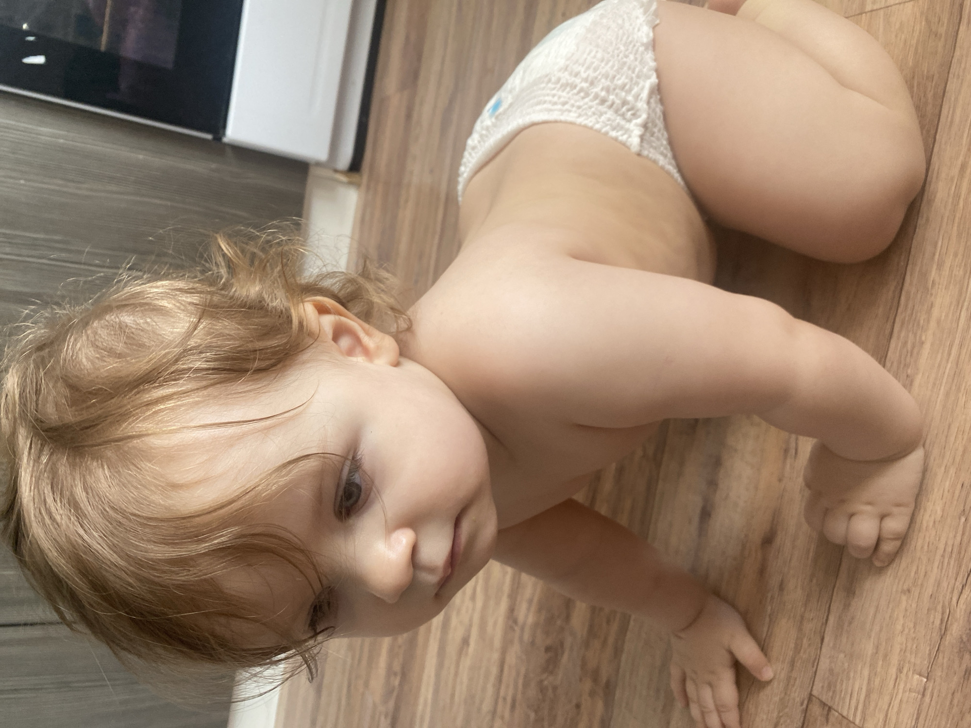A baby is captured from an elevated perspective, crawling or in a crawling position on a wooden floor. The baby is wearing a plain white diaper and has light brown or blonde, wavy hair that falls around its face. Its facial features, including light-colored eyes and a small nose, are visible as it looks downwards and slightly to its left. The baby's skin appears fair and smooth, with visible creases on its elbows and knees, indicative of its age.
In the background, the setting appears to be indoors, likely a home, with a dark-colored appliance, possibly an oven or microwave, partially visible against a white cabinet or wall on the left side of the frame. The wooden floorboards, varying in shade from light to medium brown, are a dominant feature throughout the image. The lighting is soft and even, suggesting an indoor environment during daytime. There is no discernible text visible in the image. The scene conveys a moment of typical early childhood exploration within a domestic setting in Krasnodar, Russia.