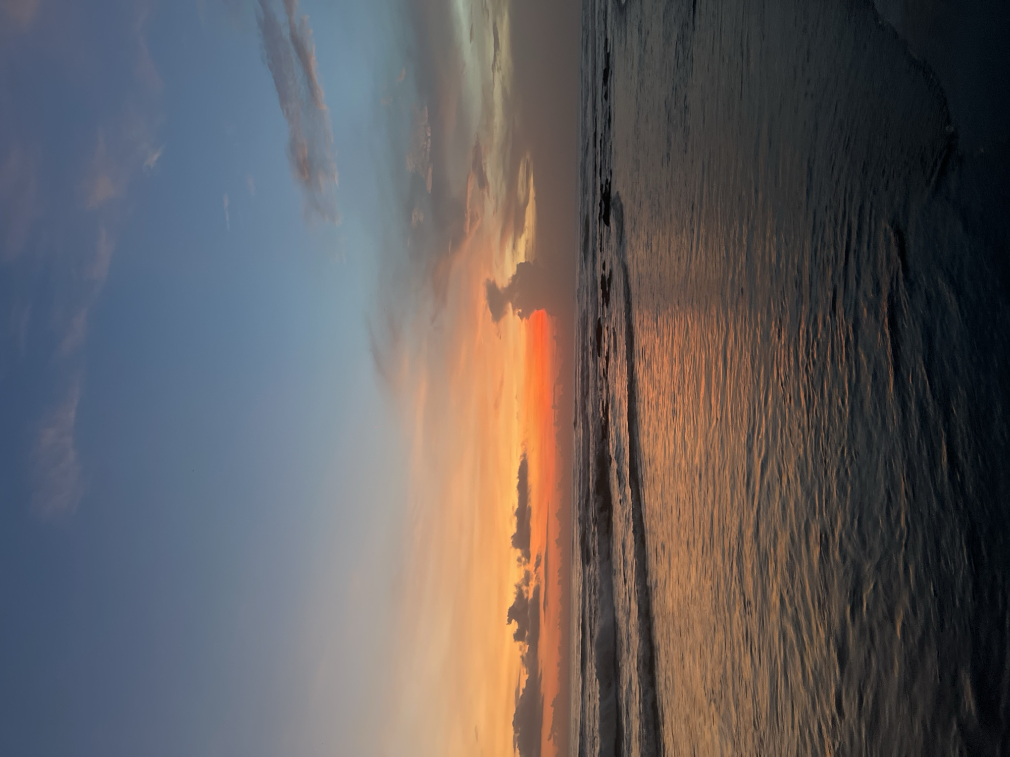 The image depicts a sunset over the ocean in Canggu, Indonesia. The sky transitions from a pale blue on the left to a vibrant orange and red in the center, with scattered clouds. The ocean on the right reflects the colors of the sunset. Small waves are visible along the shoreline. The image is taken from a low angle, possibly from the beach.