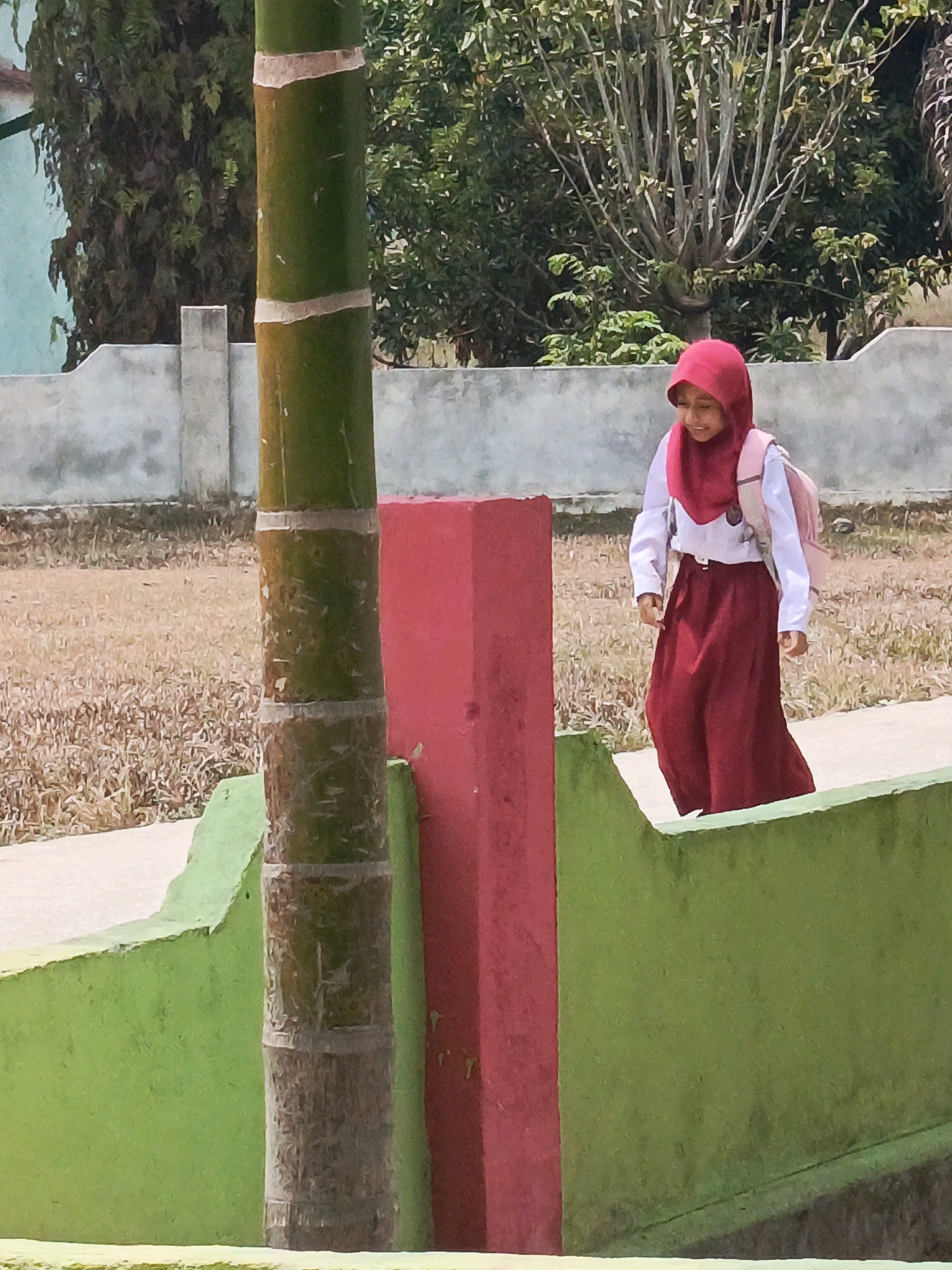 A young girl wearing a red hijab and a white shirt with a maroon skirt walks down a path next to a green and red wall. She is carrying a pink backpack. A bamboo stalk stands to the left of the girl. The background consists of dry grass, a concrete wall, and trees. The image appears to be taken outdoors in a sunny environment. The location is identified as Megang Sakti V, Indonesia.