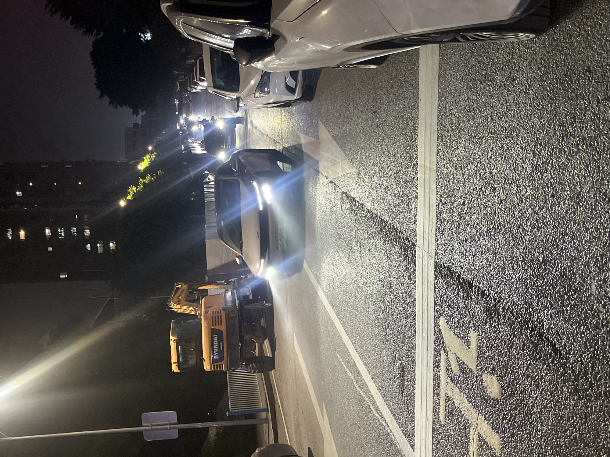 The image captures a nighttime scene on a wet asphalt road in Shapingba District, China. A bright streetlight illuminates the left side of the frame, casting a strong beam downwards. In the foreground, white lane markings, including an arrow and horizontal bars, are visible on the road surface. To the left, a large orange excavator with the brand name "HYUNDAI" emblazoned on it is parked. The excavator appears to be a construction vehicle. Several cars are present on the road; a white car is prominently positioned in the middle of the frame, facing away from the camera with its headlights on. To the right and behind this car, other vehicles are seen in traffic or parked. In the background, tall residential buildings with illuminated windows are visible against a dark sky, suggesting an urban environment. Trees can be seen on the right side of the image, partially obscuring the buildings. The overall impression is one of an active urban area at night, possibly undergoing construction or experiencing traffic.