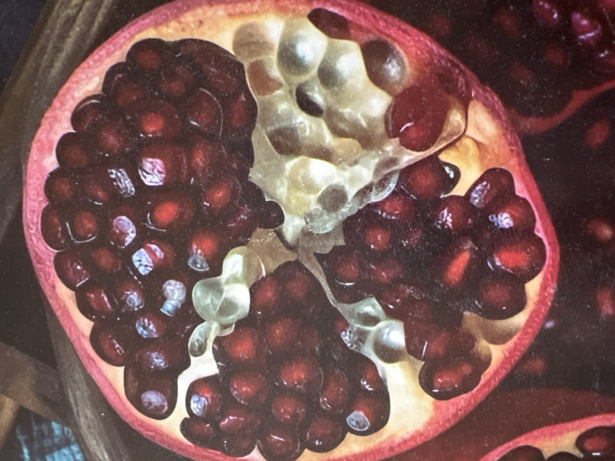 The media file is a close-up photograph of a halved pomegranate. The fruit is cut horizontally, revealing its interior filled with numerous dark red arils. The arils are clustered together, with some appearing plump and juicy, while others are slightly shriveled. White pith and membranes separate the clusters of arils. The outer rind of the pomegranate is a vibrant reddish-pink color. The lighting in the image is soft, highlighting the textures and colors of the fruit. The background is dark and out of focus, with hints of wood grain suggesting a natural or rustic setting. There is no visible text in the image. The scene appears to be a still life, focusing solely on the pomegranate. The location context of Minsk, Belarus, does not provide any specific visual cues within the image itself.