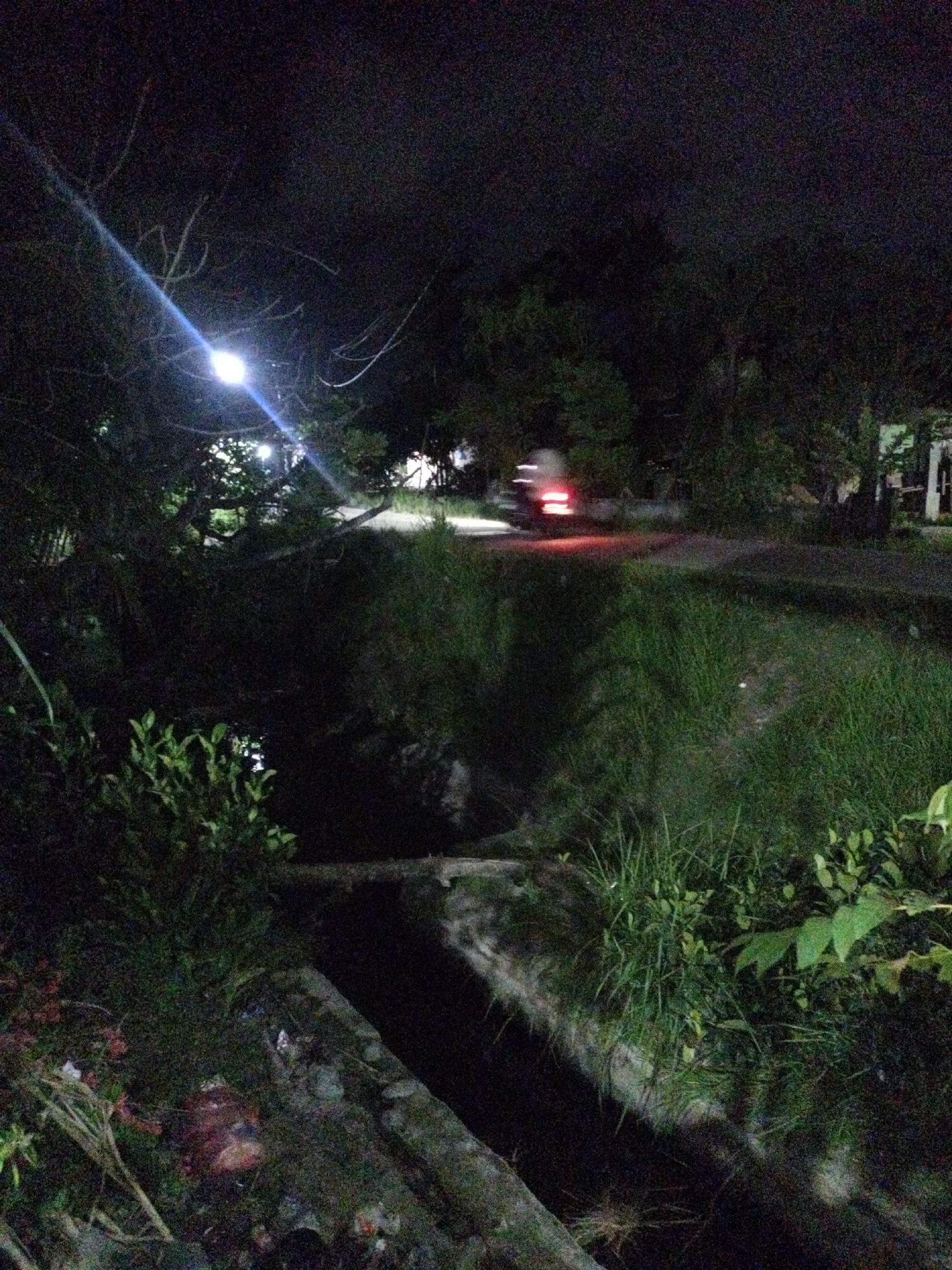 A nighttime scene in Seuneuboklhong, Indonesia. A narrow concrete channel with stagnant dark water is in the foreground, flanked by overgrown grass and foliage. A street lamp illuminates the left side of the frame, casting a bright glow. A motorcycle with its taillights visible is traveling on a paved road in the middle ground, disappearing into the darkness of trees in the background. A house structure is visible on the right of the frame, adding to the scene’s setting in a neighborhood. The scene is dimly lit, with the majority of the area shrouded in darkness.