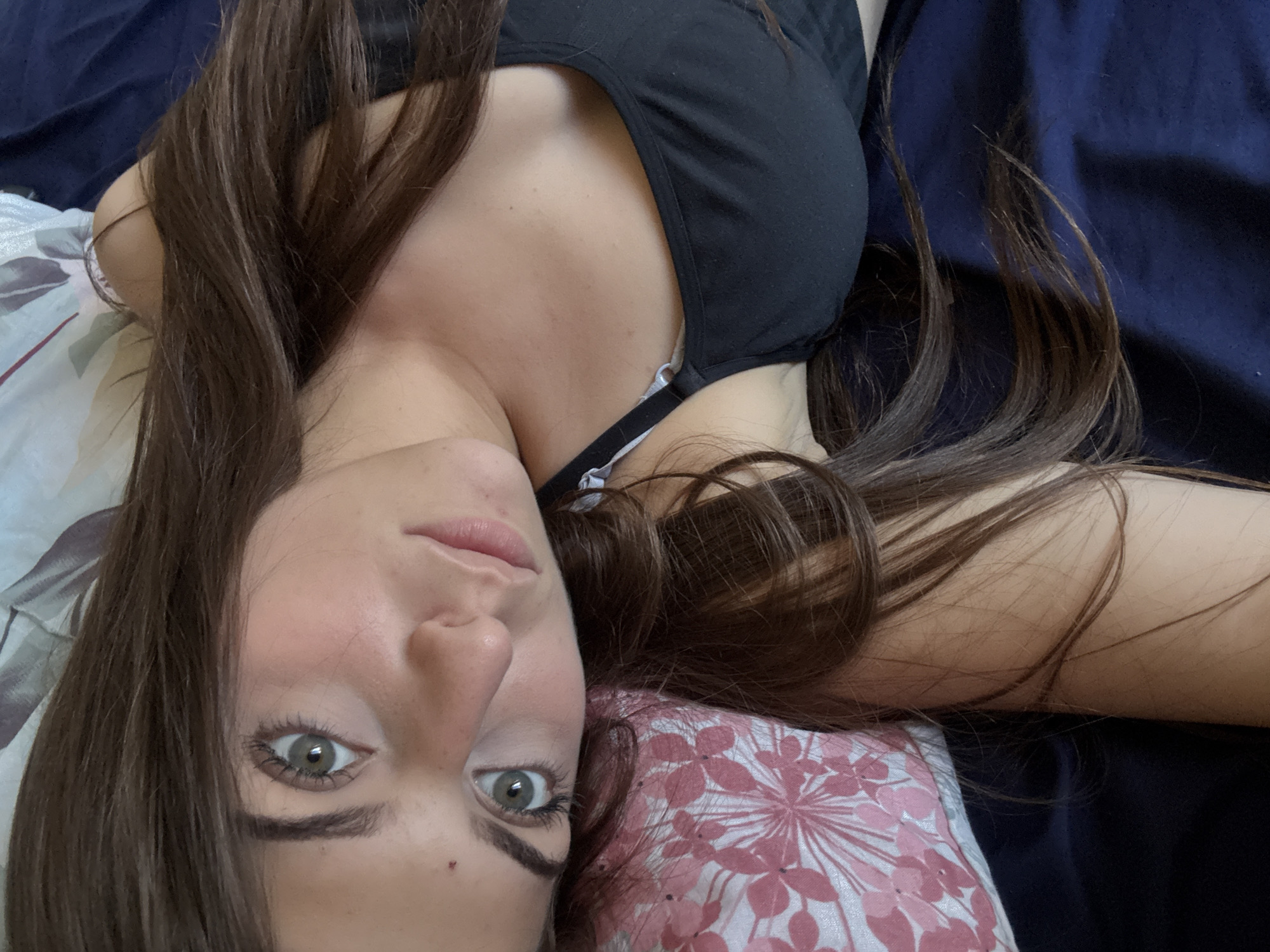 A woman with fair skin and long brown hair is lying on her back. Her head rests on a pillow with a pink and white floral pattern. She is wearing a black top with thin straps. The background includes a blue, presumably bed-covering, material. The location is not discernible based on the visual data in the image.