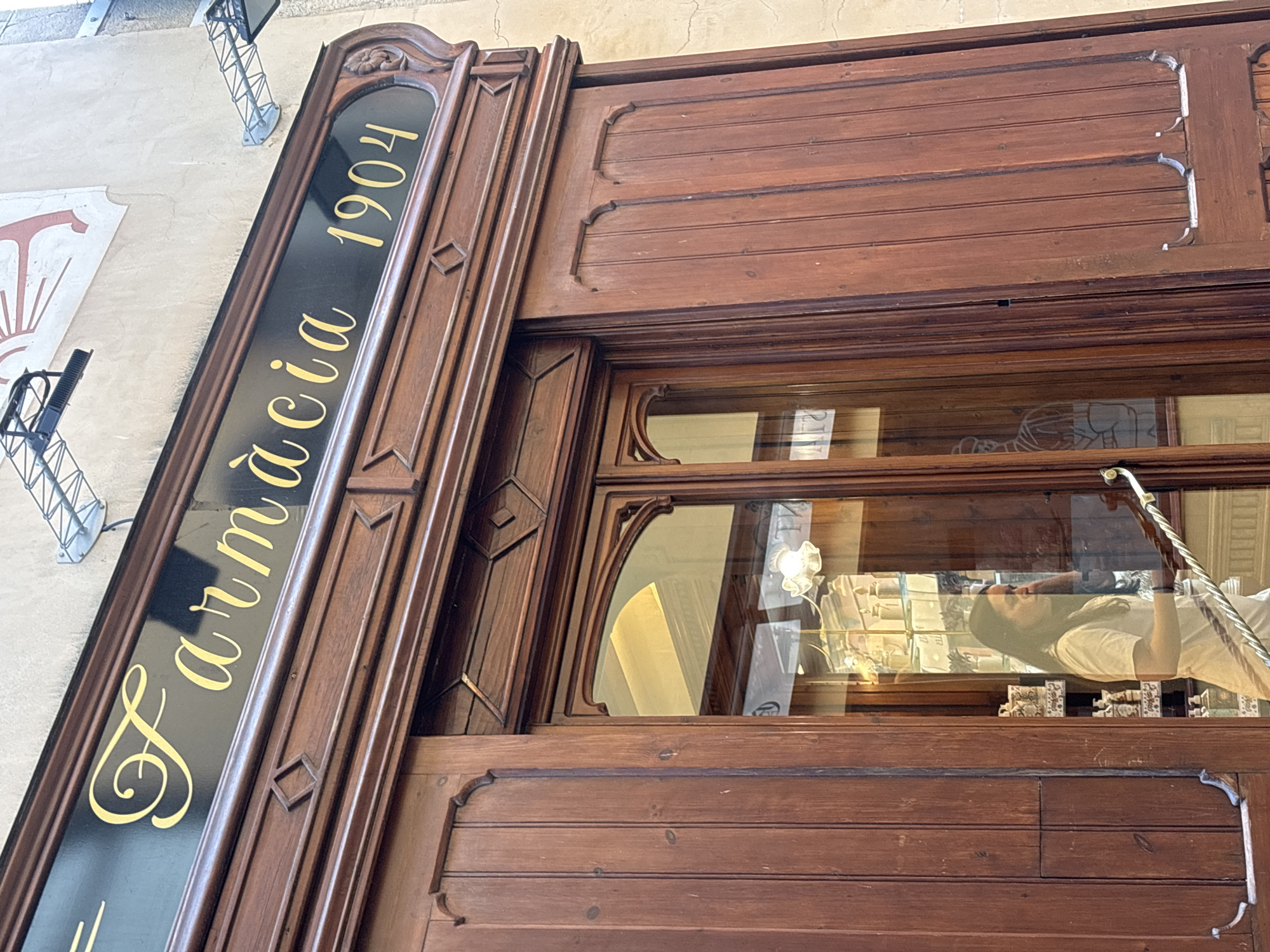 The image shows the exterior of a pharmacy named "Farmàcia 1904" in Palafrugell, Spain. The facade is made of dark wood with ornate carvings. Above the entrance, a black sign with gold lettering reads "Farmàcia 1904". The entrance features double wooden doors, one of which is slightly ajar, revealing a reflection in the glass. A woman wearing a white t-shirt is visible through the glass, holding a cane or a similar object. Inside, shelves are stocked with various products, suggesting the interior of a traditional pharmacy. The building's exterior wall is light beige, and there are metal fixtures attached to the wall above the sign, possibly for lighting or decorative purposes. The overall impression is of an old, established business.