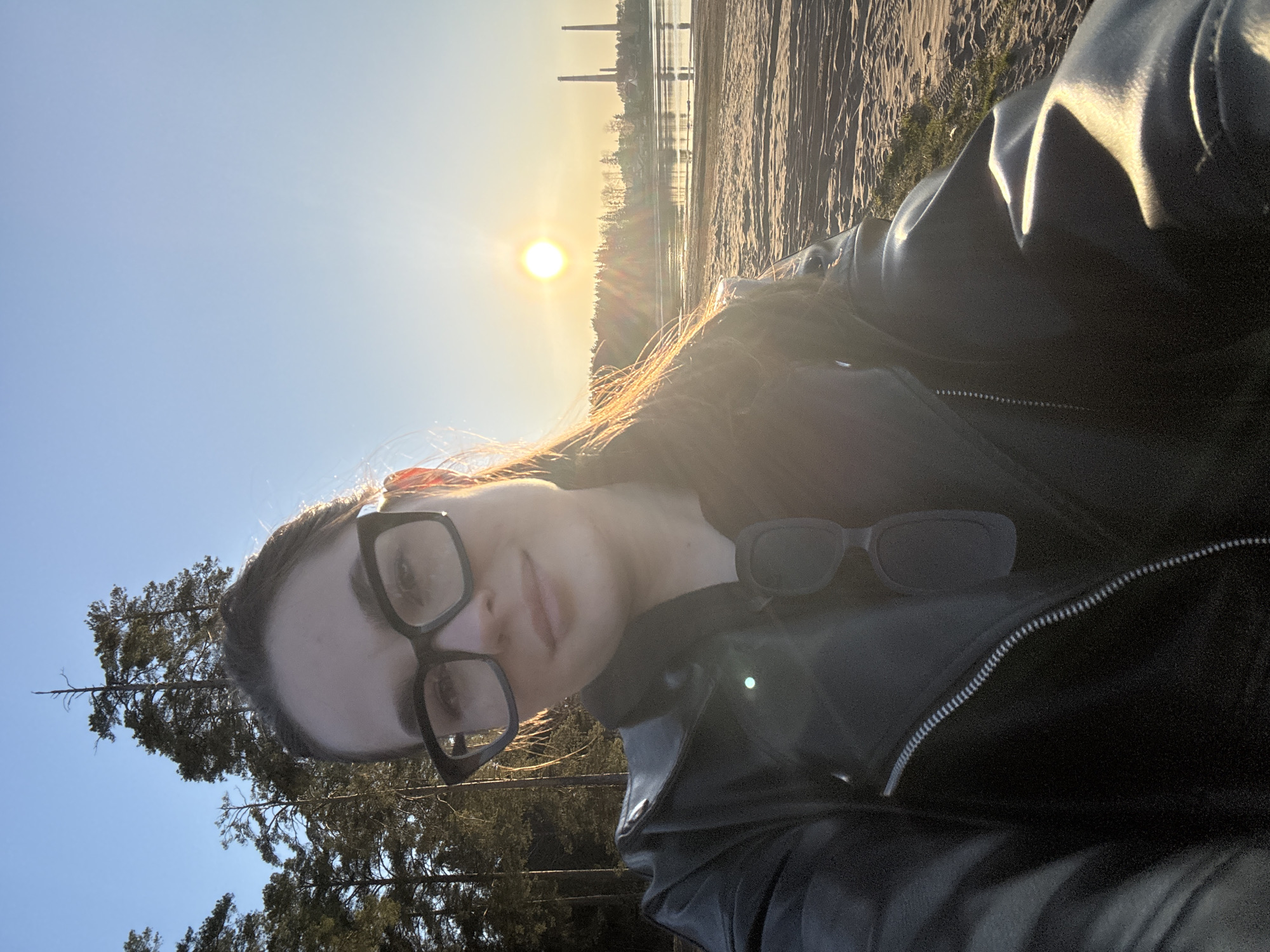 The photograph is a selfie taken outdoors at sunset, likely in Chaikovsky, Russia, given the mention of the city. A woman with brown hair and fair skin wears large, black-framed eyeglasses and a black leather jacket. The sun is visible in the background, positioned over a body of water with a sandy shoreline. Structures resembling industrial chimneys are also present in the background on the horizon. Trees are visible on the left side of the frame against a blue sky. Sunglasses are partially visible near the woman's jacket. The photograph is rotated, placing the sky and trees on the left and the shoreline at the top.