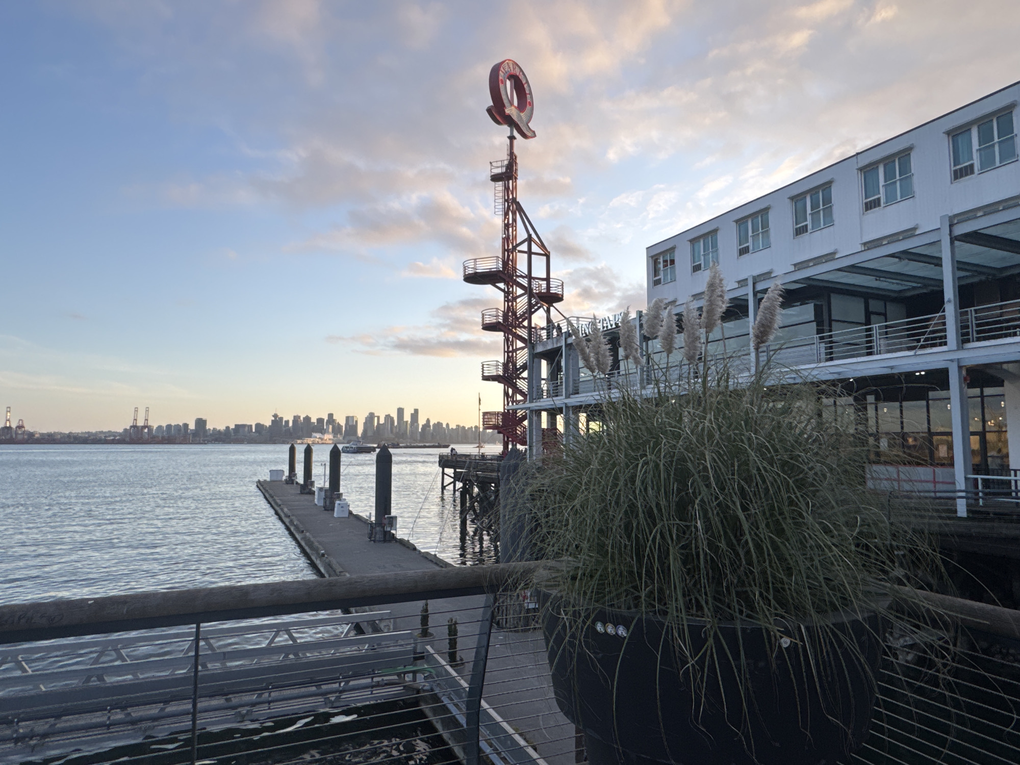 The image captures a waterfront scene in North Vancouver, Canada, likely at Lonsdale Quay. A long pier stretches into the water, with several black cylindrical bollards and smaller white equipment boxes visible. In the background, the Vancouver skyline is visible.
On the right side of the frame, there's a multi-story white building with dark windows. A red metal tower with a large, red "Q" on top stands next to the building. In the foreground, a large black planter with tall grass obscures part of the scene. The railing of a walkway is also visible.
The sky is lightly cloudy and the lighting suggests it could be dawn or dusk. No people are distinctly identifiable in the image.