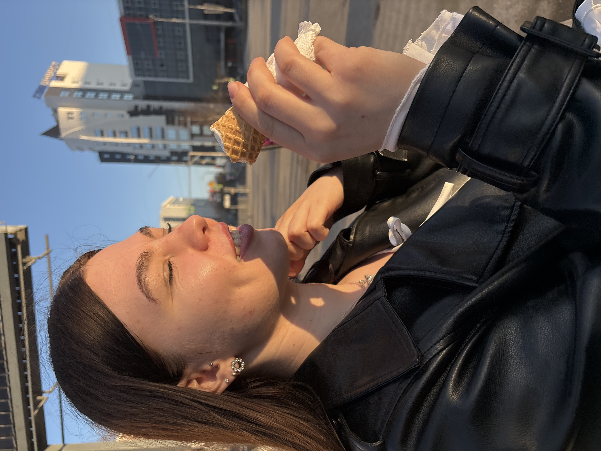 A young woman is eating an ice cream cone outdoors in Perm, Russia. She is holding the cone in her right hand, which is wrapped in a paper napkin. She has her eyes closed as she licks the ice cream. The woman is wearing a black leather jacket over a white blouse with frilly sleeves. She has dark hair and several piercings in her left ear. In the background are modern buildings, a street, and a blue sky.