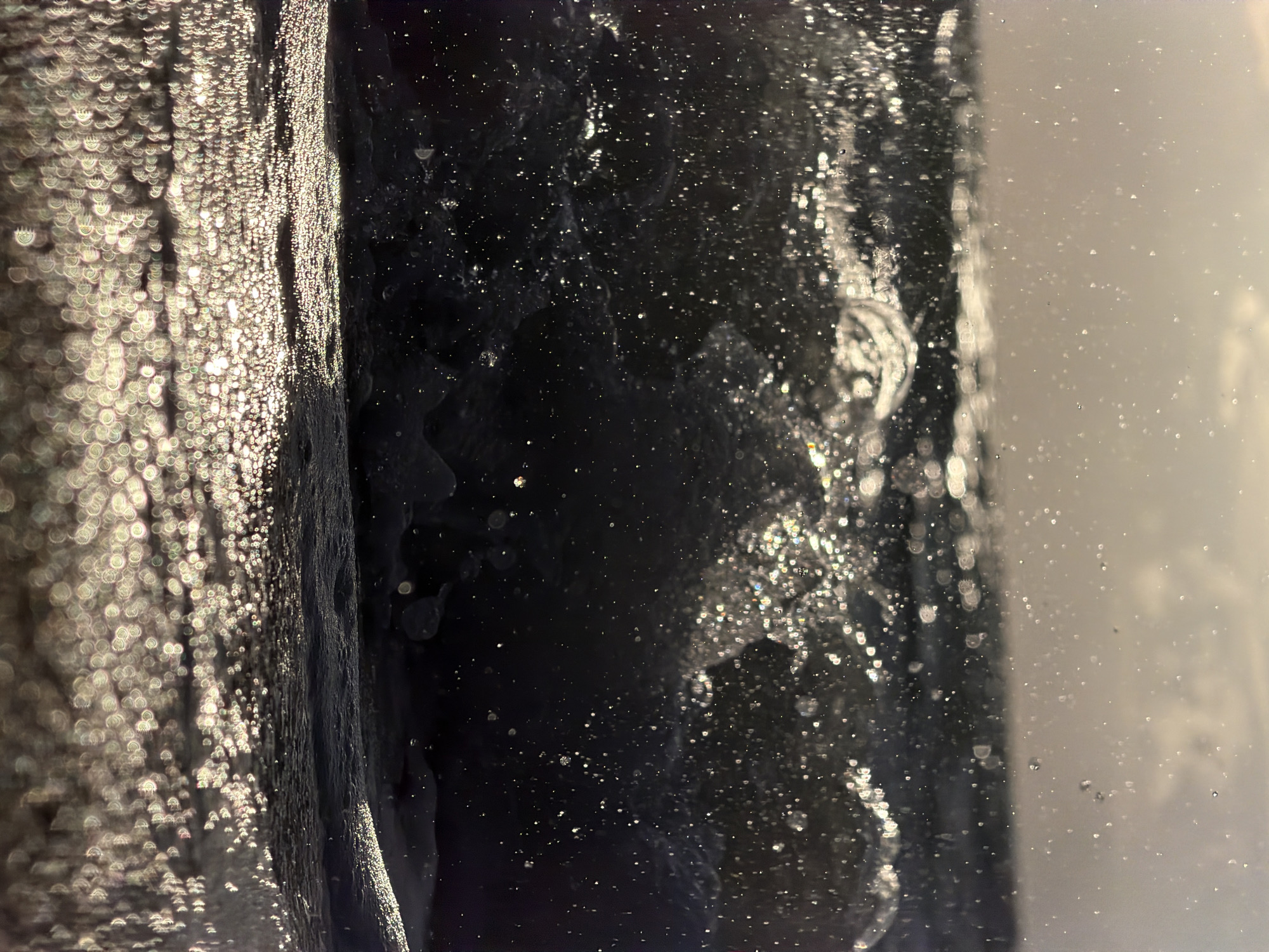The image shows a close-up, low-angle shot of a breaking wave at sea. The water is dark and turbulent, with many small droplets and splashes caught in the air, illuminated by a bright light source, likely the sun, creating a shimmering effect. The foreground is filled with blurred, bokeh-like circles of light reflecting on the wet sand or shallow water. The background is a hazy, overcast sky with some indication of clouds. The overall impression is one of dynamic movement and the play of light on water. This scene is consistent with a beach on the island of Ko Samui, Thailand, known for its tropical waters and natural beauty.