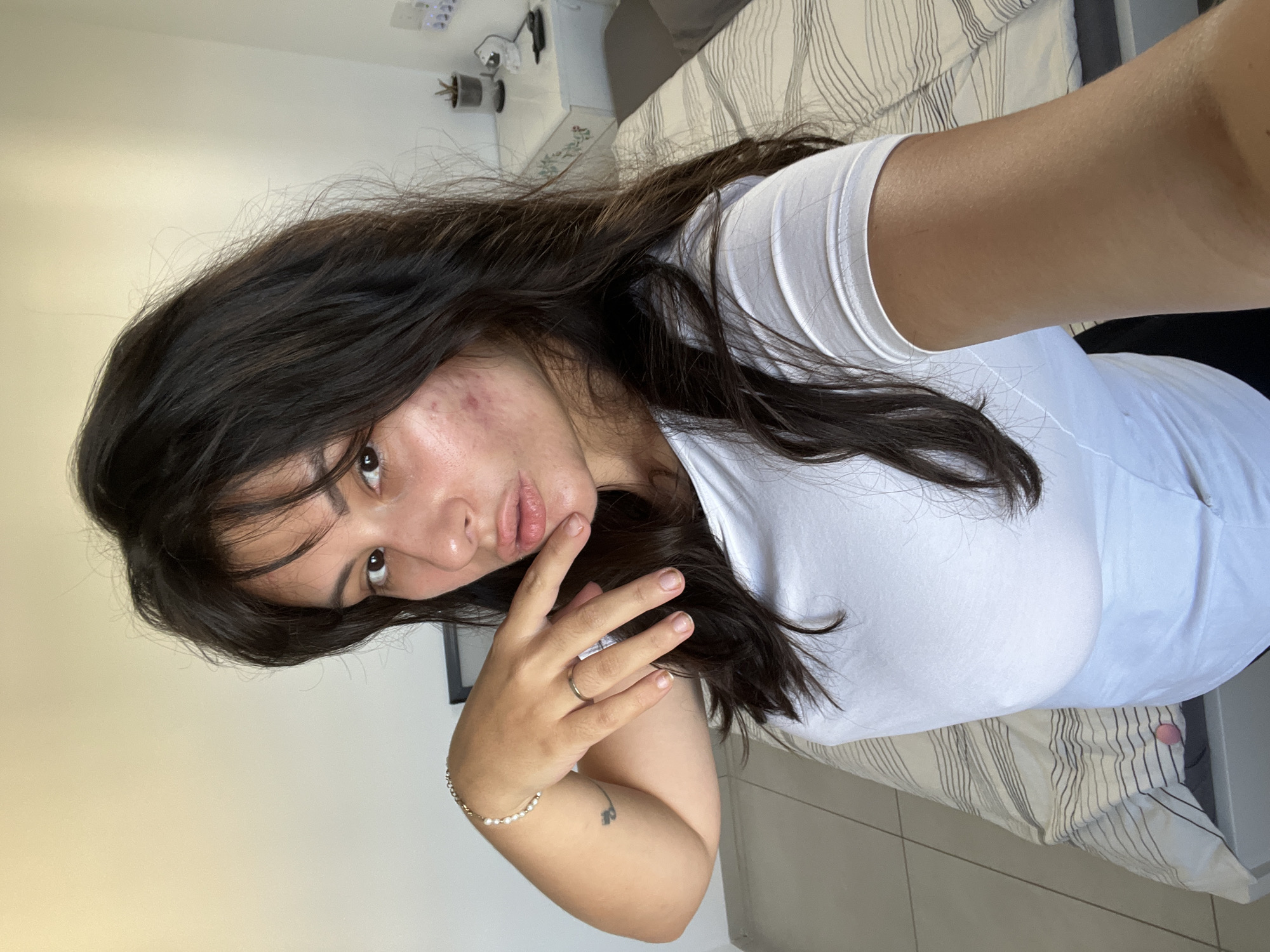 A young woman with long, dark, wavy hair is taking a selfie in what appears to be a bedroom. She is wearing a white t-shirt. Her left arm is bent, with her hand near her face. She has a bracelet on her left wrist and a ring on her left ring finger. There are blemishes on her cheek. In the background, a bed with striped bedding is visible, along with a white dresser and a tiled floor. The lighting suggests it is daytime. The scene appears to be indoors. The location is specified as Kiryat Bialik, Israel.