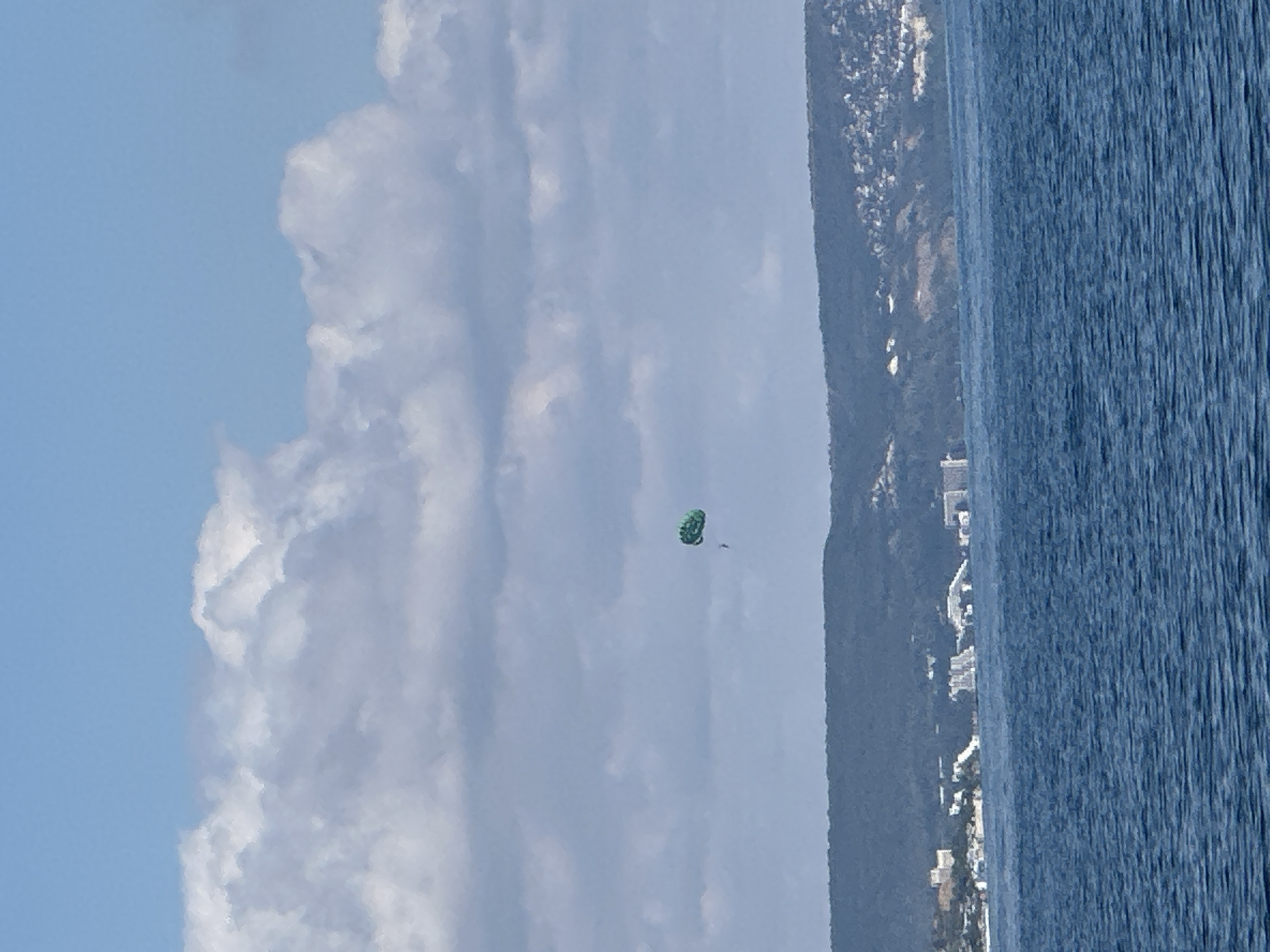 A parasailer is suspended in the air over a body of water, attached to a green parachute. The parasailer is a small figure in the distance. The water is a deep blue with rippling waves. In the background, a lush green hillside with scattered buildings and a coastline is visible. The sky above is a pale blue with scattered white clouds. The scene appears to be a scenic view of a coastal resort area.