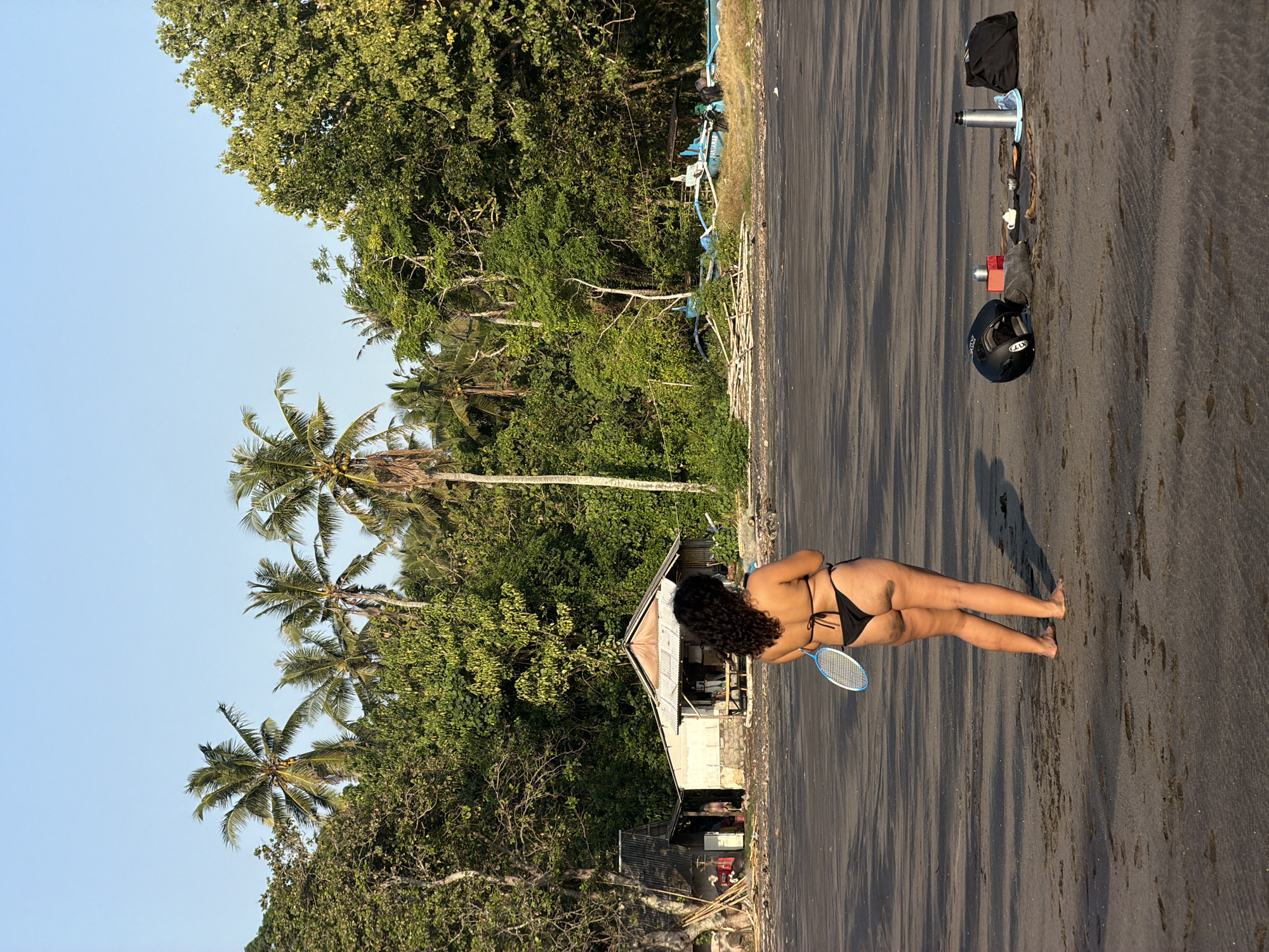 The image depicts a tropical coastal scene in Selemadeg, Indonesia, with a lone individual on a dark sand beach facing away from the camera. A woman with dark, curly hair, wearing a black two-piece bikini, stands barefoot, holding a blue and white badminton racket in her left hand. To her right on the sand, a collection of items is arranged: a black motorcycle helmet, a small red rectangular container, a white teapot with accompanying small cups on a dark mat, a silver thermos, a blue plate or frisbee, and a black duffel bag.
The background features dense, vibrant green tropical vegetation, including numerous tall palm trees with visible coconuts. Towards the left, two simple, rustic structures with corrugated metal roofs are visible amongst the foliage. Further to the right, partially obscured by trees, are at least two light blue traditional outrigger fishing boats (jukung) on the sand, accompanied by piles of cut wood or bamboo. The sky is clear and light blue. The beach exhibits ripple patterns and scattered footprints in the dark sand. The lighting suggests daytime, with soft shadows cast to the right.