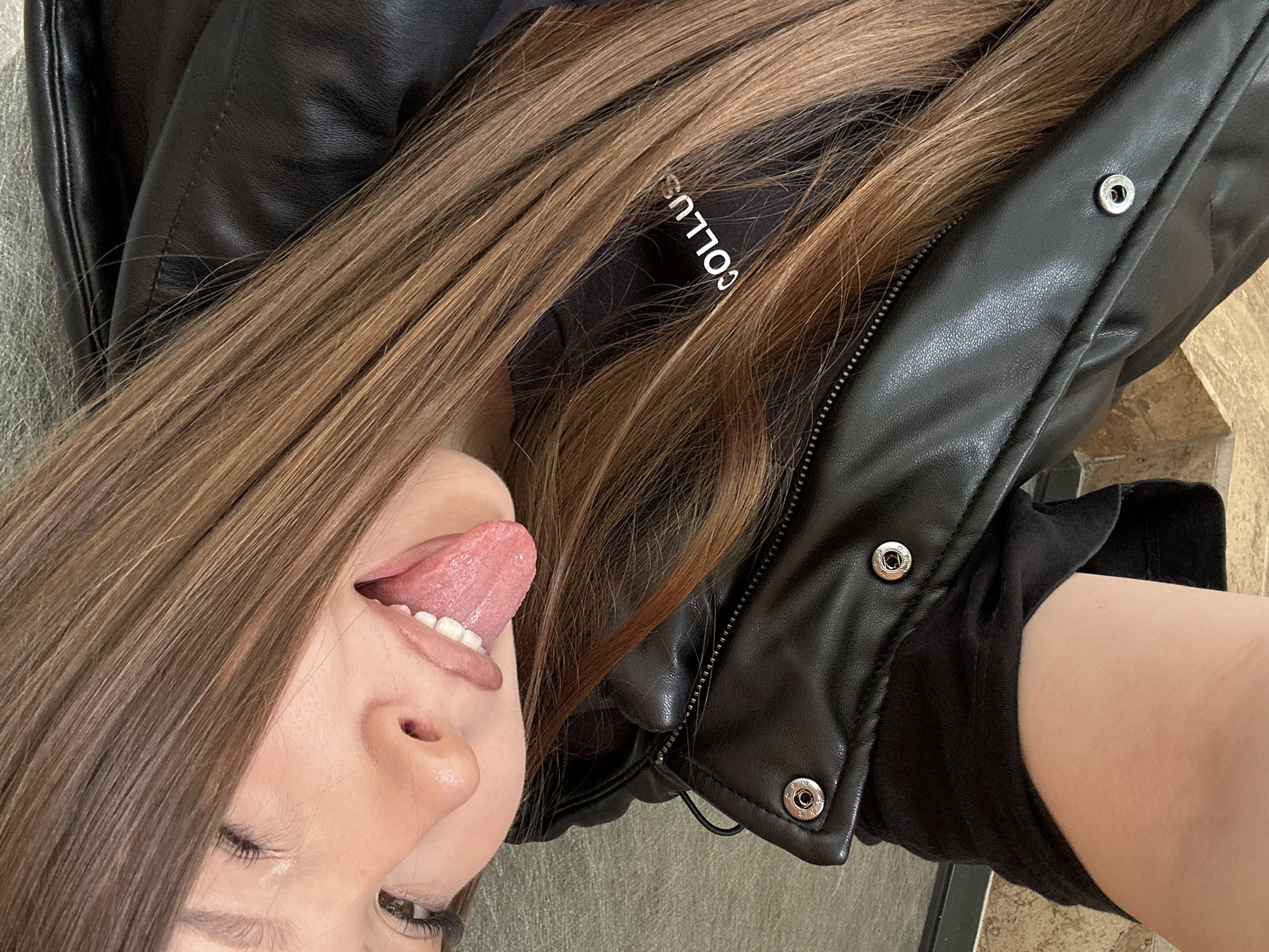 A close-up, angled shot shows a fair-skinned person with long, light brown hair lying down, facing left and sticking their tongue out. They are wearing a black leather-like jacket with silver snap buttons and a zipper visible. A black shirt is also visible beneath the jacket. The background appears to be an indoor setting with a light brown tile floor. There are no explicit indications that the image was taken in Varna, Bulgaria.