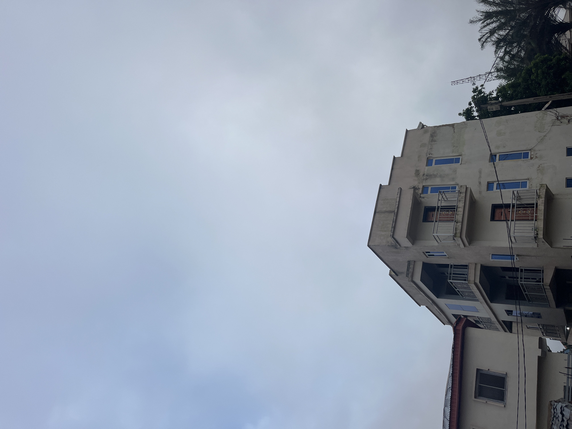 This image captures a three-story building in Jos, Nigeria, under a cloudy sky. The building's facade is a muted beige, showing signs of wear and tear. Each floor features balconies with metal railings, and several rectangular windows with blue-tinted glass are visible. A smaller, adjacent building with a red corrugated metal roof is partially visible to the left. In the background, a transmission tower and lush green palm trees suggest an urban or suburban setting. The overall impression is one of a functional, perhaps older, residential or commercial structure in Jos.