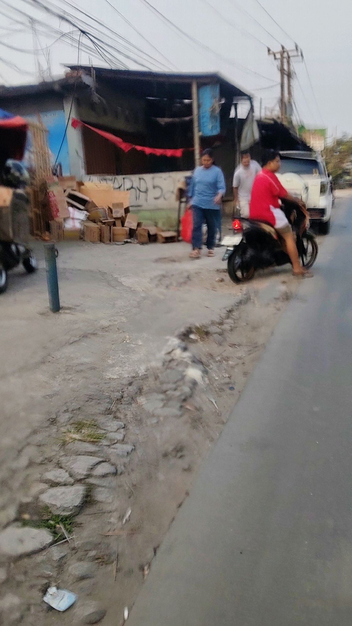 In Mekarmukti, Indonesia, an overcast day features a roadside scene with a simple, weathered structure and individuals engaged in activity. A woman in a blue shirt and jeans walks with a red bag in her hand, accompanied by a man in a white shirt behind her. To the right, a person in a red shirt and white shorts rides a black motorcycle, facing away from the camera. The structure, possibly a stall or shop, has a dark, corrugated roof with some red fabric hanging from it. Stacks of cardboard boxes are visible near the structure. The ground is a rough, dirt-like surface with scattered rocks and patches of grass. A blue pole stands near the left edge of the image. A white van is parked in the background. The overall impression is one of a simple, everyday scene in a developing urban environment.