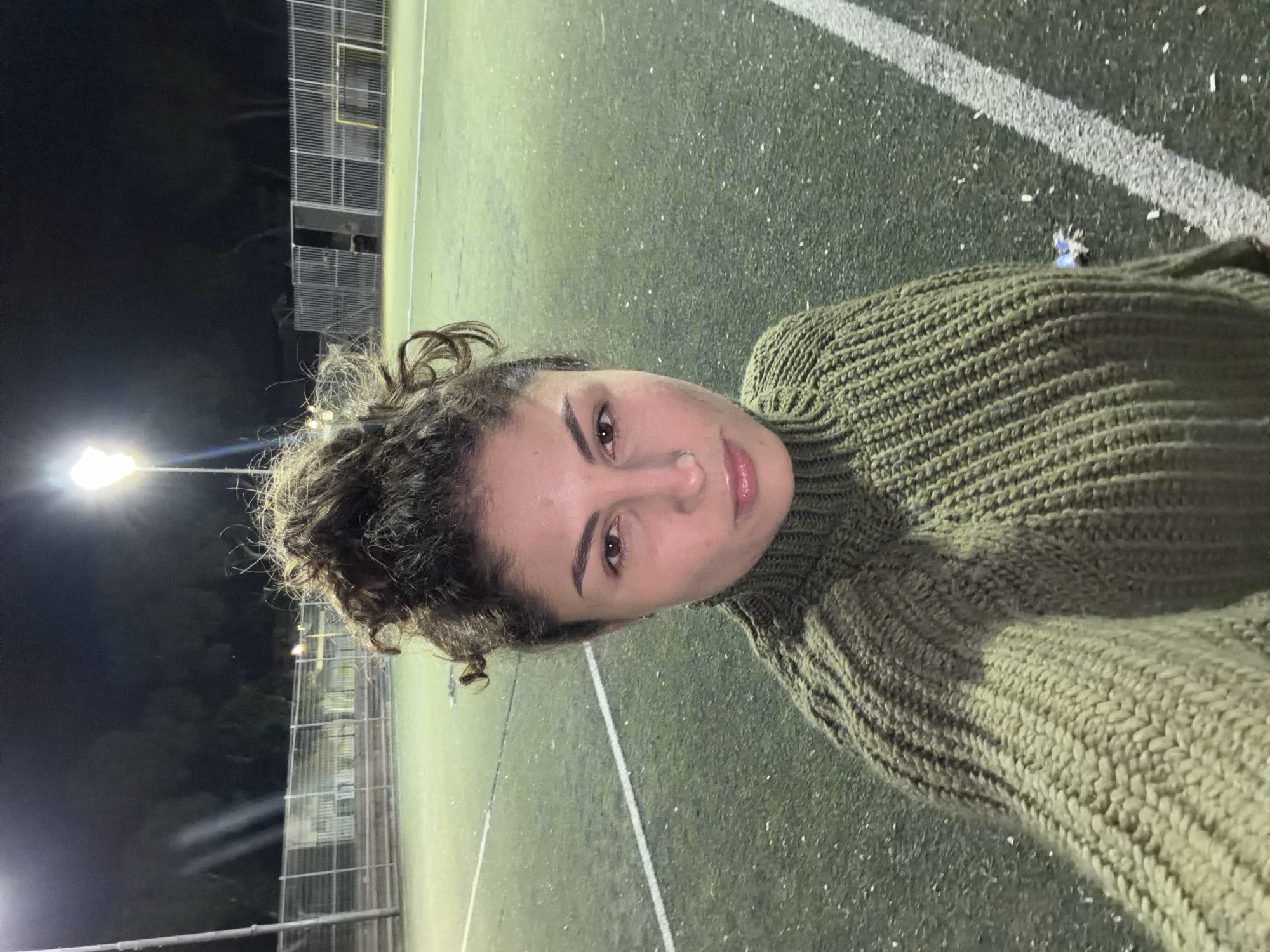 The media file features a portrait-oriented selfie of a young woman taken at night on an artificial turf sports field in Kiryat Bialik, Israel. The woman is positioned on the right side of the frame, looking directly at the camera with a neutral expression. She has curly brown hair, partially styled in an updo, and is wearing a thick, olive-green knitted turtleneck sweater.
The background consists of a green artificial turf field, clearly marked with white lines, typical of a sports pitch. Numerous small white specks, likely rubber infill material, are scattered across the artificial grass. A small blue object is visible on the ground near the edge of the woman's sweater.
The scene is illuminated by bright artificial lighting, with a prominent light source visible on the left side, casting a glow across the dark sky. A tall, wire-mesh fence extends across the mid-ground, suggesting the field is enclosed. The overall setting indicates an outdoor sports facility at night. No discernible text is visible within the image.