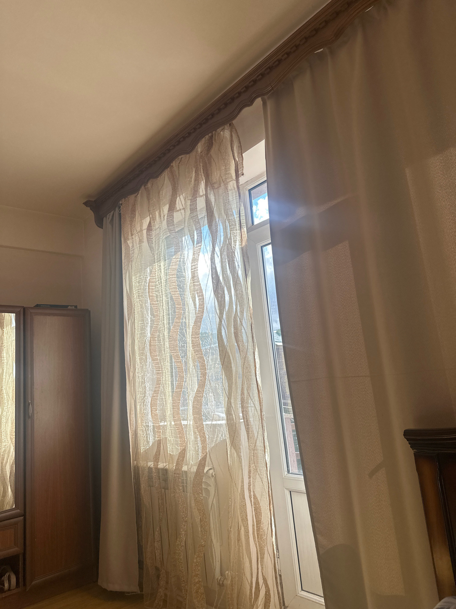 A bedroom interior is shown with natural light coming from a window. On the left, a tall, dark wood wardrobe with a mirrored door is partially visible. In the center, a window is covered by two layers of curtains: an inner sheer curtain with a wavy pattern in beige and cream, and an outer opaque curtain in a light beige color. The outer curtain is pulled to the right, revealing a sliver of the window. The window frame is white. Sunlight streams through the sheer curtain, creating a bright, hazy effect. A white radiator is visible behind the sheer curtain. The ceiling is light beige, with a dark wood decorative trim along the top of the window area. The flooring appears to be a light wood laminate. The overall impression is a well-lit room, likely in a residential setting in Gyumri, Armenia, given the context. No people are present in the image.