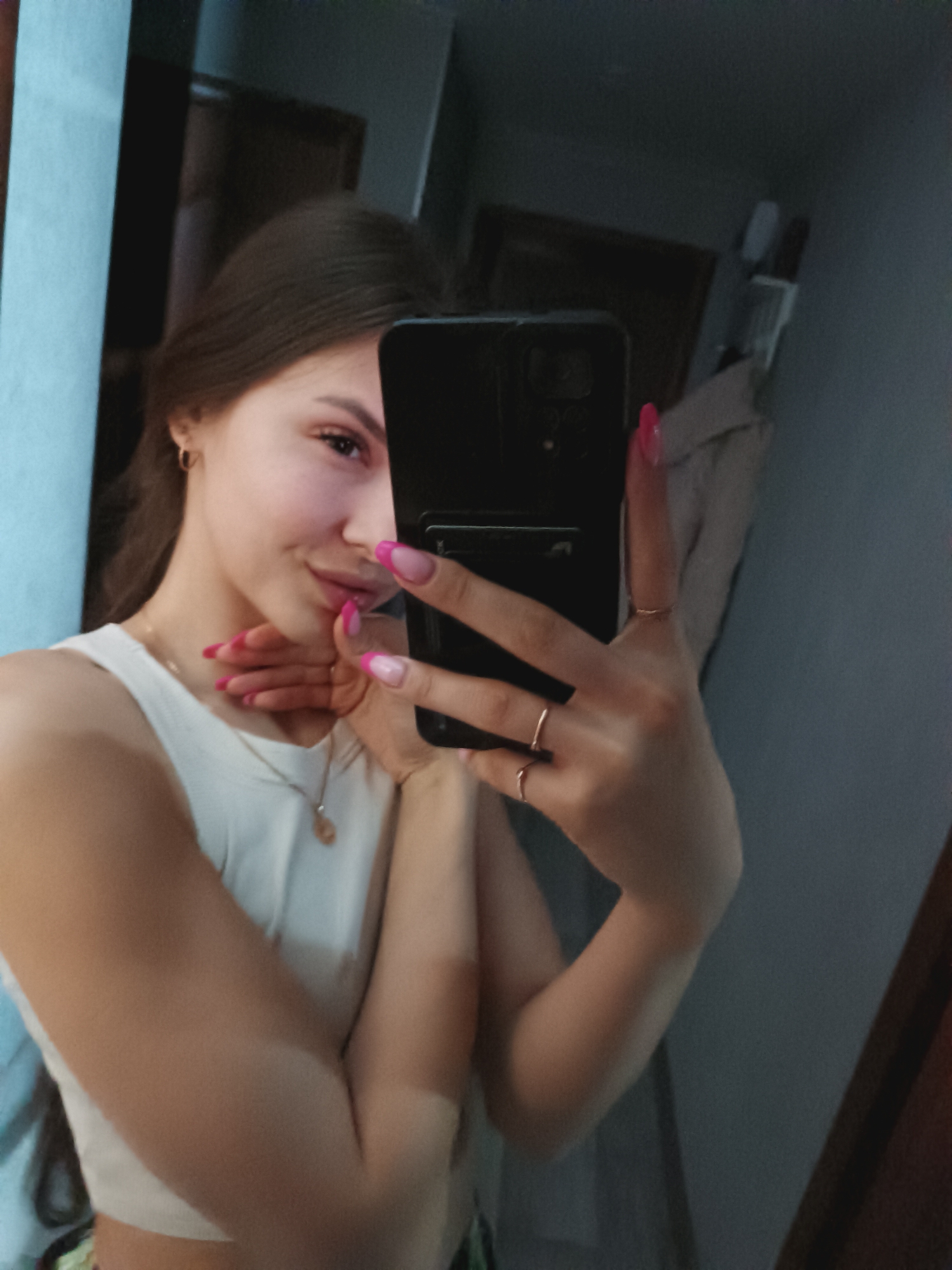 In the city of Unknown, Unknown, a female subject is depicted taking a mirror selfie indoors. The subject, with medium-length brown hair, is visible from the chest up, wearing a white sleeveless crop top. A gold-colored necklace with a small pendant is around her neck, and a small gold hoop earring is visible in her left ear. Her fingernails are manicured with a pink French tip design. She holds a black smartphone vertically with her right hand, partially obscuring the right side of her face. Three rings are visible on her right hand: one on the ring finger, one on the middle finger, and one on the index finger. Her left hand is positioned near her chin and neck. Her visible left eye is open, and she exhibits a faint smile. The background, reflected in the mirror, shows an interior space with a light blue or teal wall on the left. A dark wooden door frame or door is visible on the right side of the reflection, and further right, a faint light-colored object, possibly a garment on a hanger, is against a dark grey wall. The lighting in the scene appears subdued.