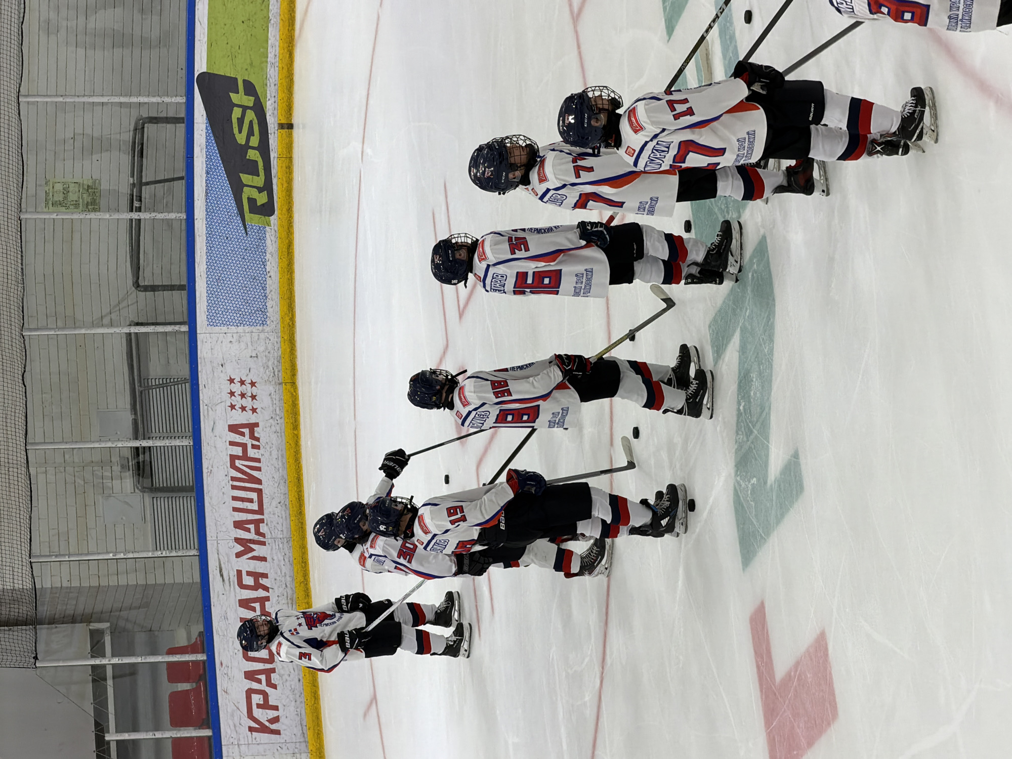 The image shows a group of young hockey players lined up on an ice rink. They are wearing hockey jerseys with numbers and logos. The players have hockey sticks and are wearing helmets. There are advertisements and markings on the rink. The location is possibly in Almetyevsk, Russia based on the text "КРАСНАЯ МАШИНА" and other Russian text visible.